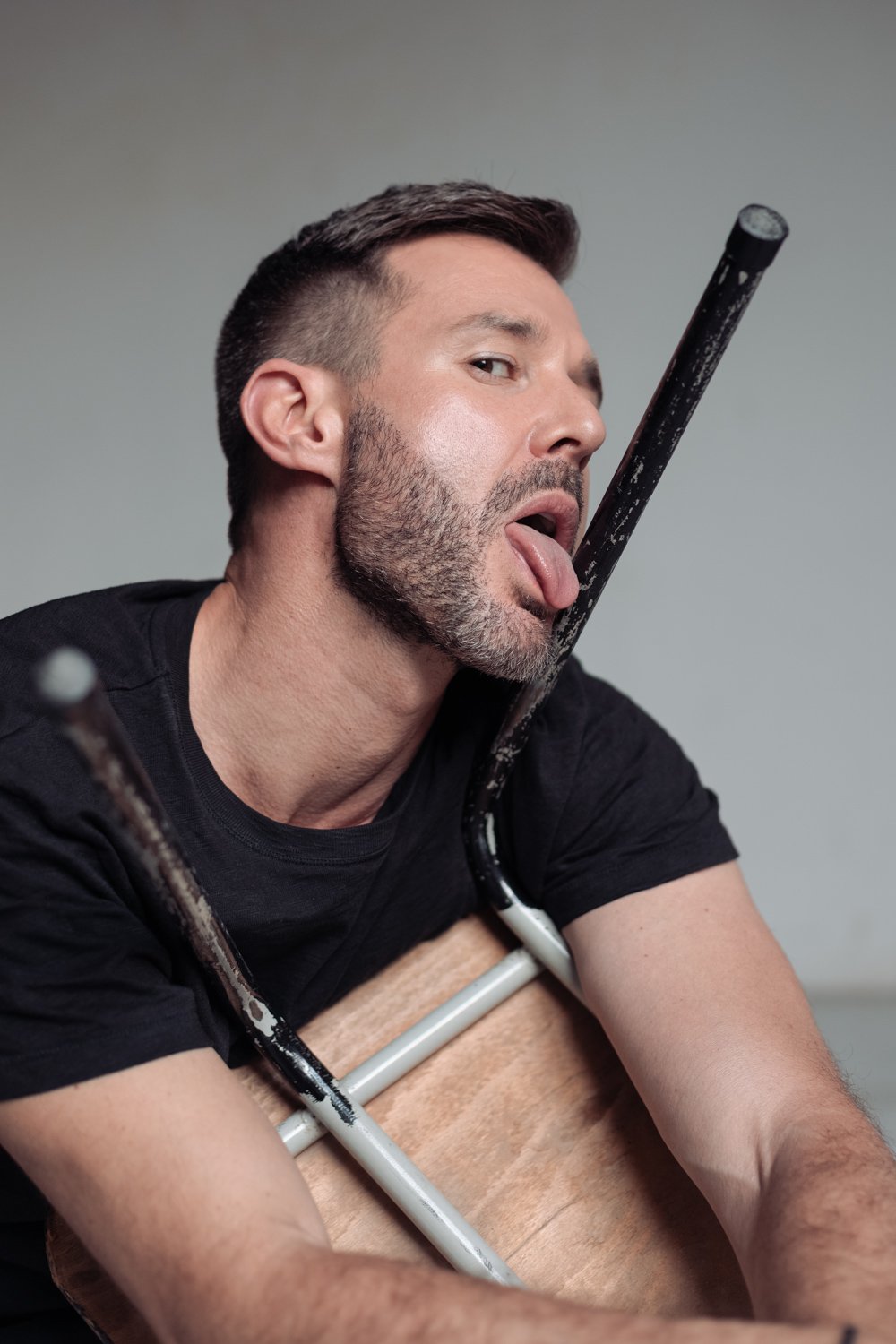 A man with short dark hair and a beard is posing with a chair, sticking out his tongue and looking at the camera. He is wearing a black t-shirt and is leaning on a wooden table or chair. The background is plain and gray.