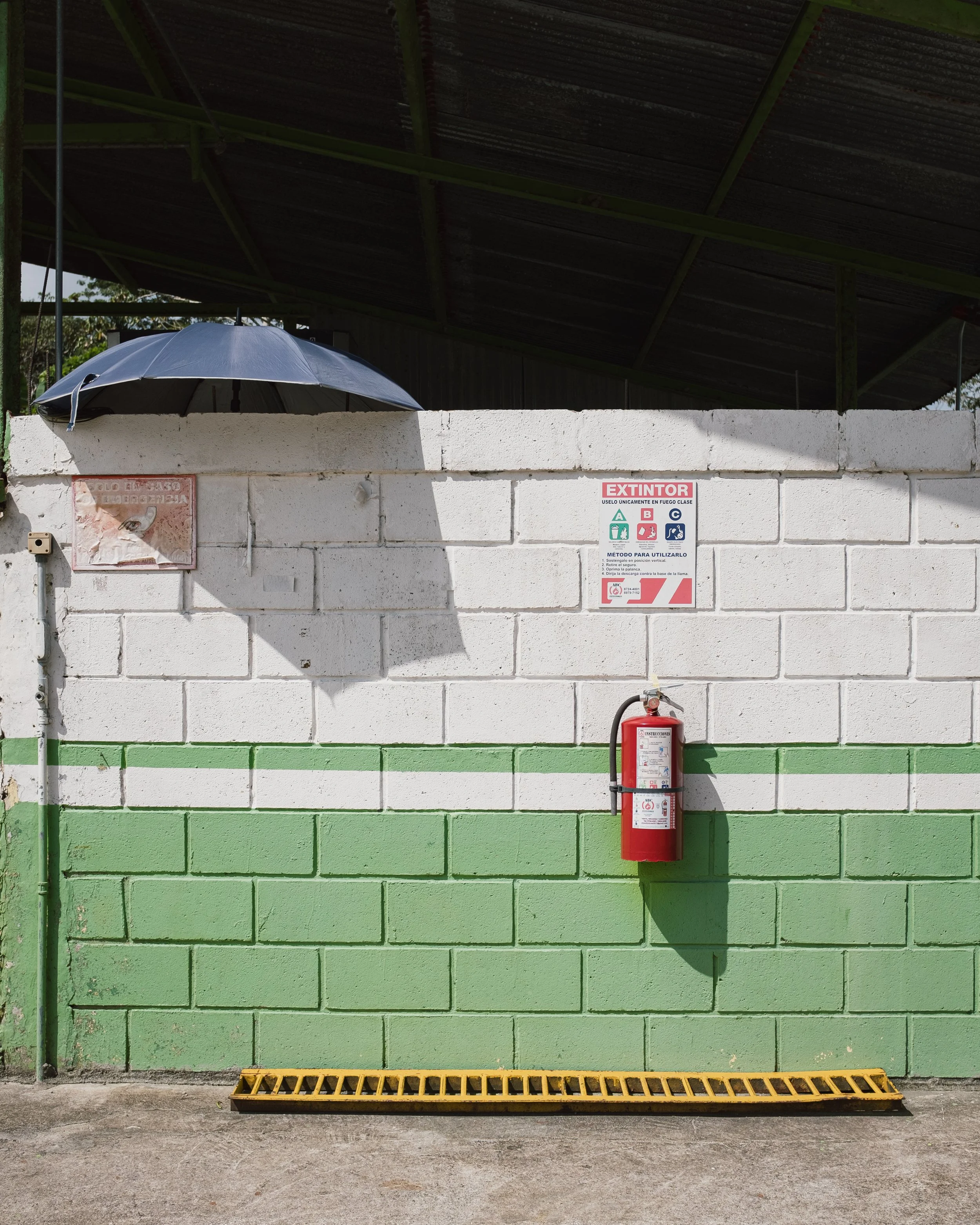 A white brick wall with a green lower section, a fire extinguisher mounted on it, a warning sign, and an umbrella casting a shadow. There's also a yellow drain grate on the ground in front.