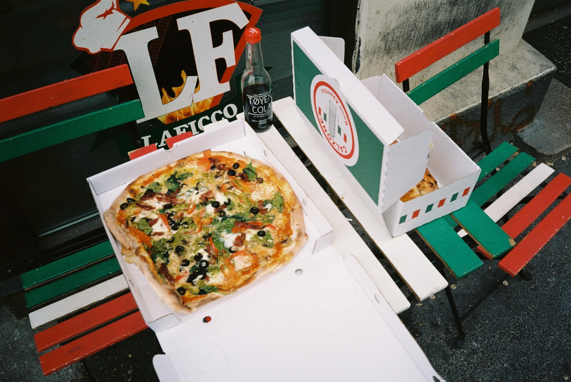 A pizza with vegetables in an open pizza box on a bench outside. Next to it, another closed pizza box and a bottle of cola. There are red, white, and green painted slats on the bench.