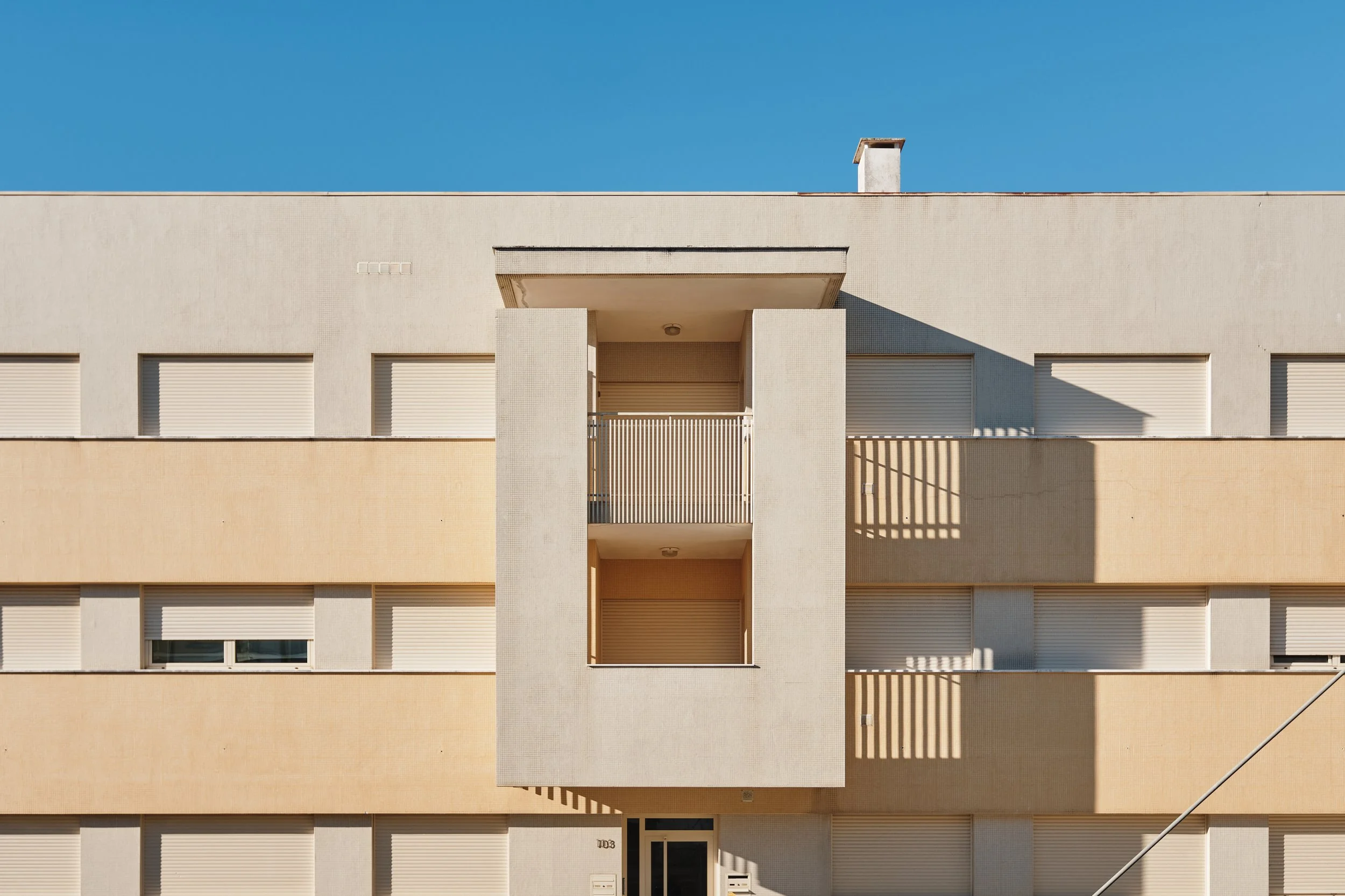 Front view of a modern apartment building with beige and white exterior walls, two balconies with white railings, and closed shutters on the windows, set against a clear blue sky.