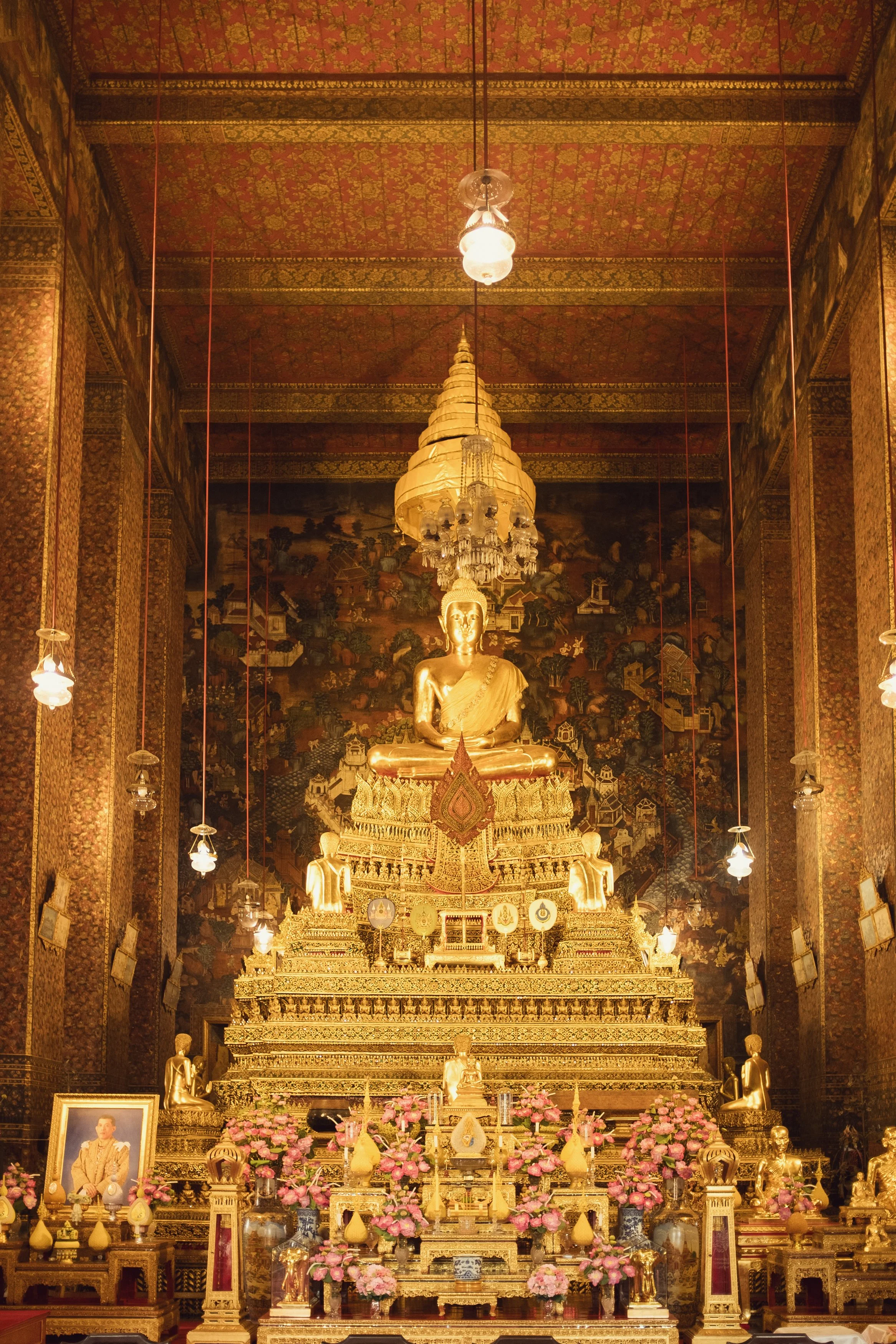 Golden Buddha statue in a richly decorated temple with intricate wall murals, hanging lanterns, and floral arrangements.