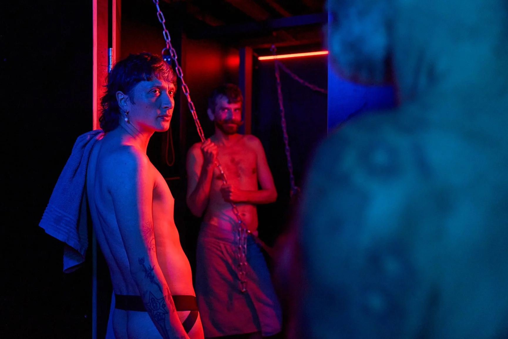 Three people in a dimly lit room with red and blue lighting, two of whom are shirtless and appear to be chained or connected by chains; one man with tattoos on arm looking away, and a man with a beard standing behind, holding a chain. A partially visible person on the right is out of focus.