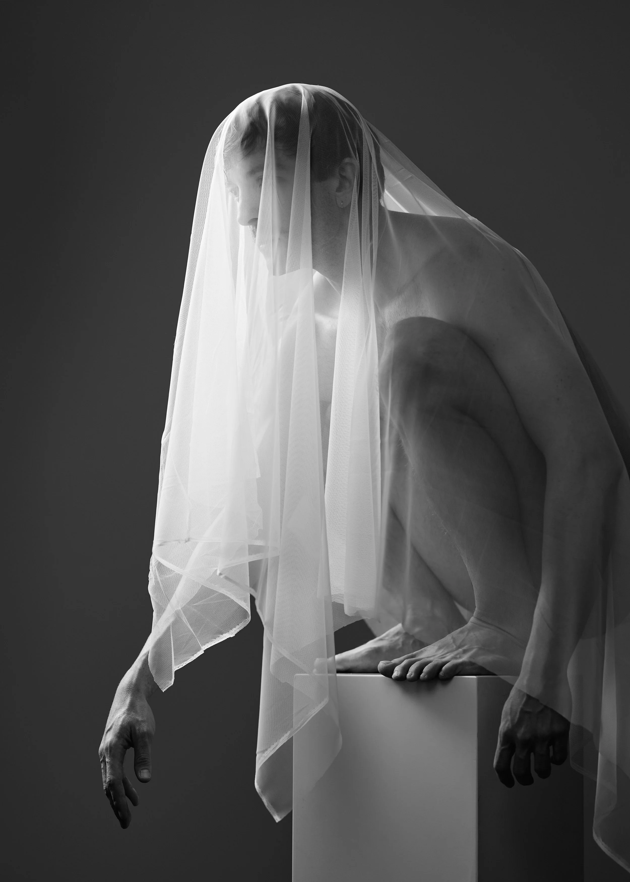 A person wearing a veil, is crouched on a white pedestal with hands resting on the sides, looking downwards, in a black and white studio setting.
