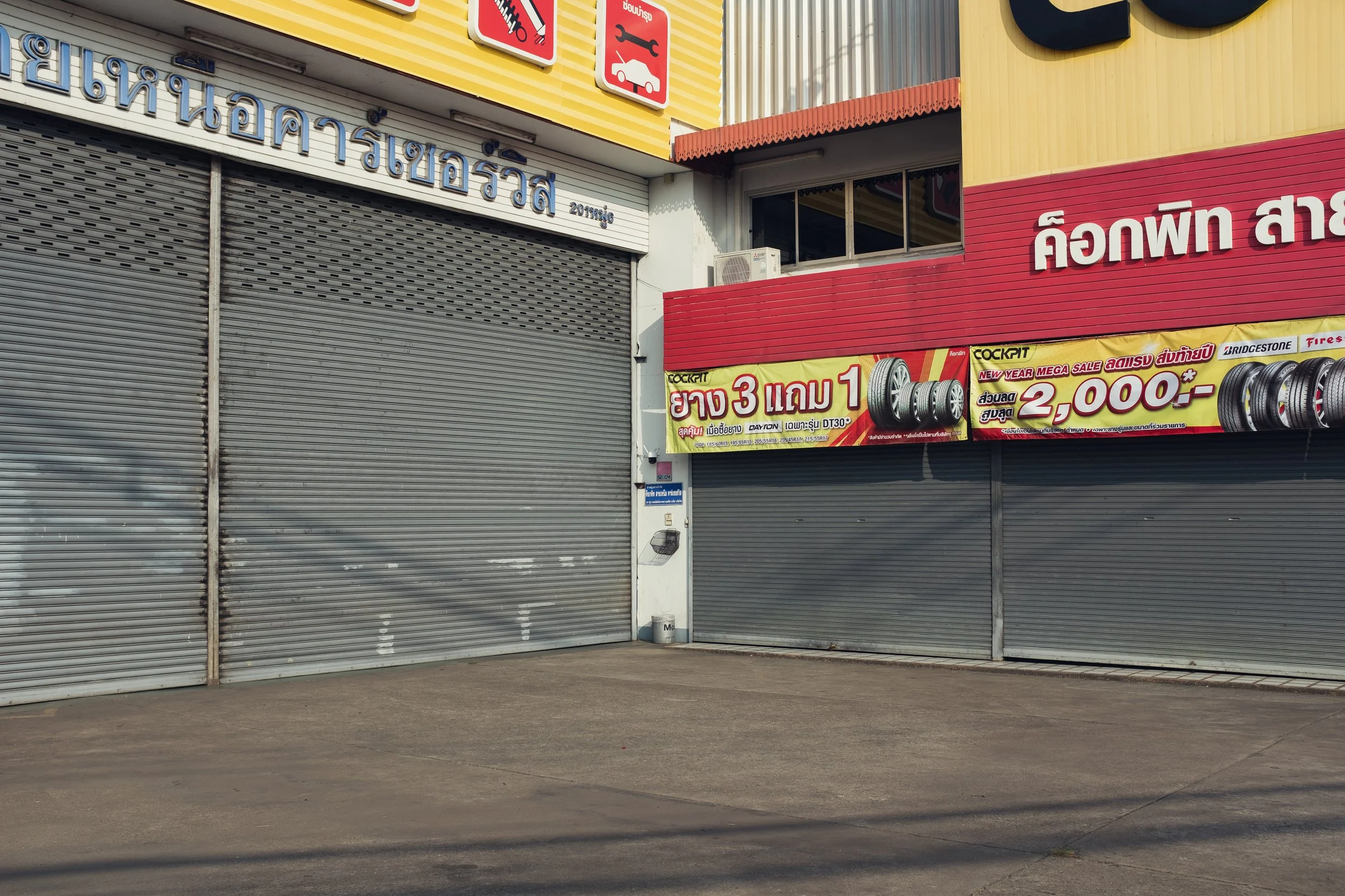 Closed metal storefront shutters with a colorful sign in Thai language advertising tire discounts in an outdoor shopping area.