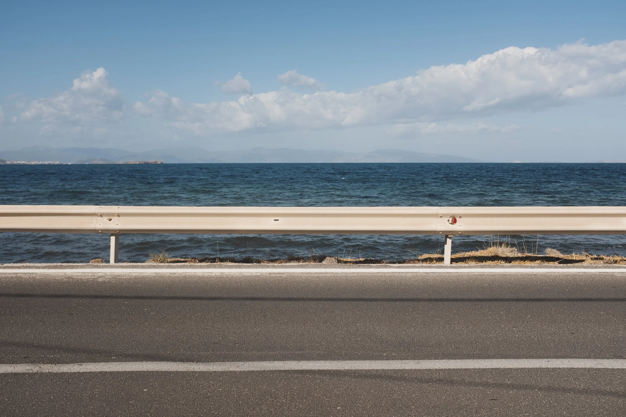 View of the ocean with waves, a clear blue sky with scattered clouds, a metal guardrail, and part of a paved road.