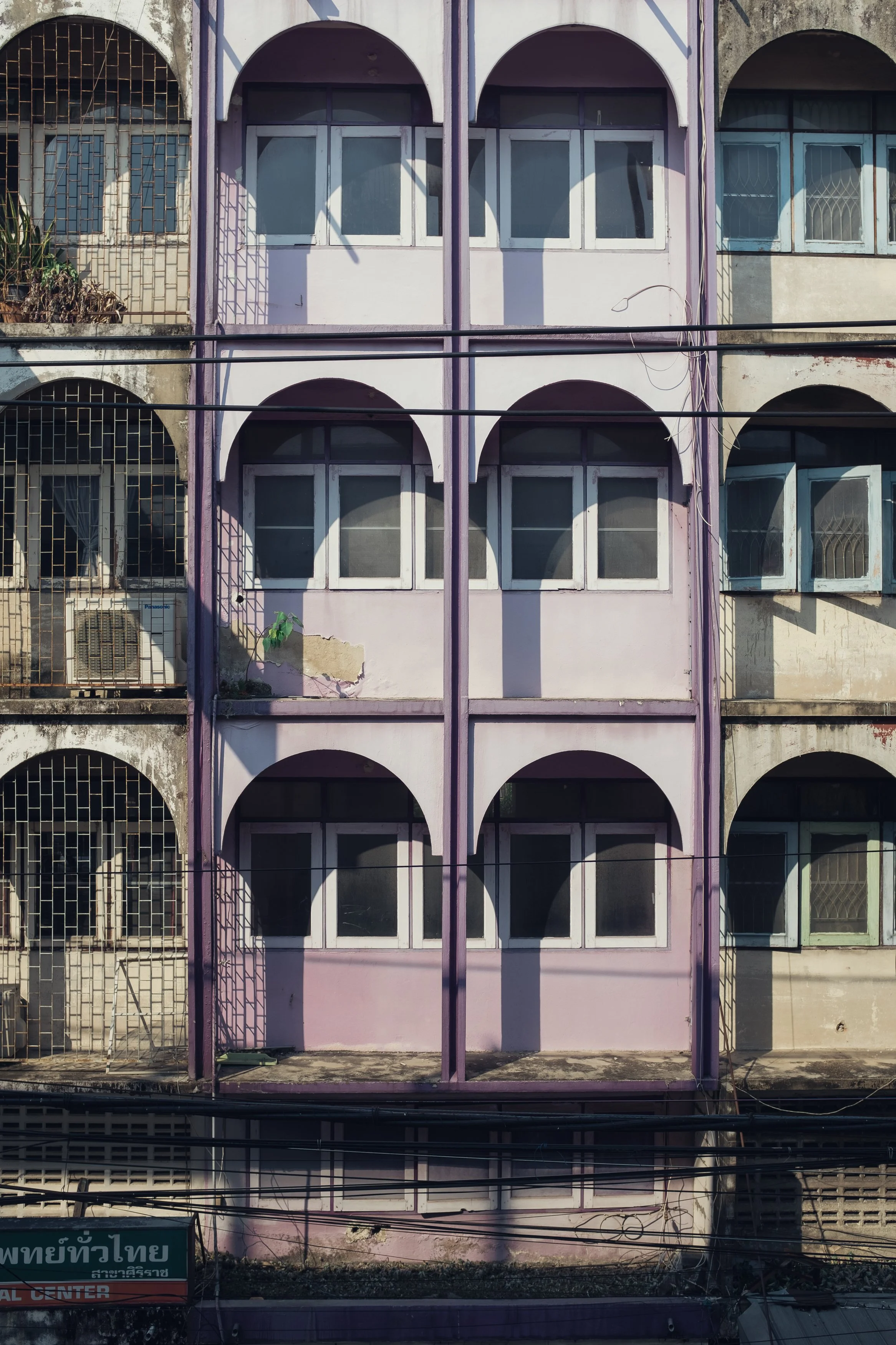 Front view of a worn-out multi-story building with pink and purple walls, featuring arched windows and metal bars on some windows, and visible electrical wires across the facade.