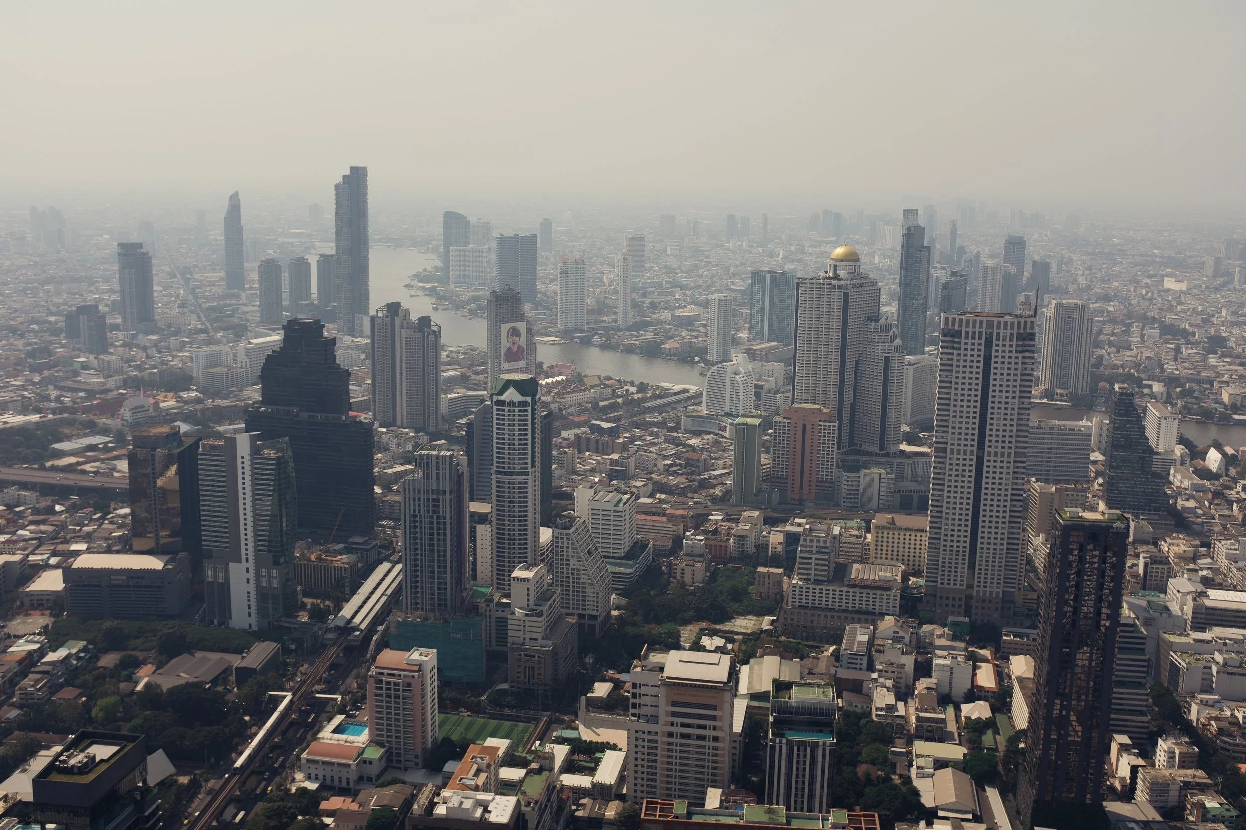 Aerial view of a city skyline with numerous tall skyscrapers, a river running through, and a mix of modern and older buildings, under a hazy sky.