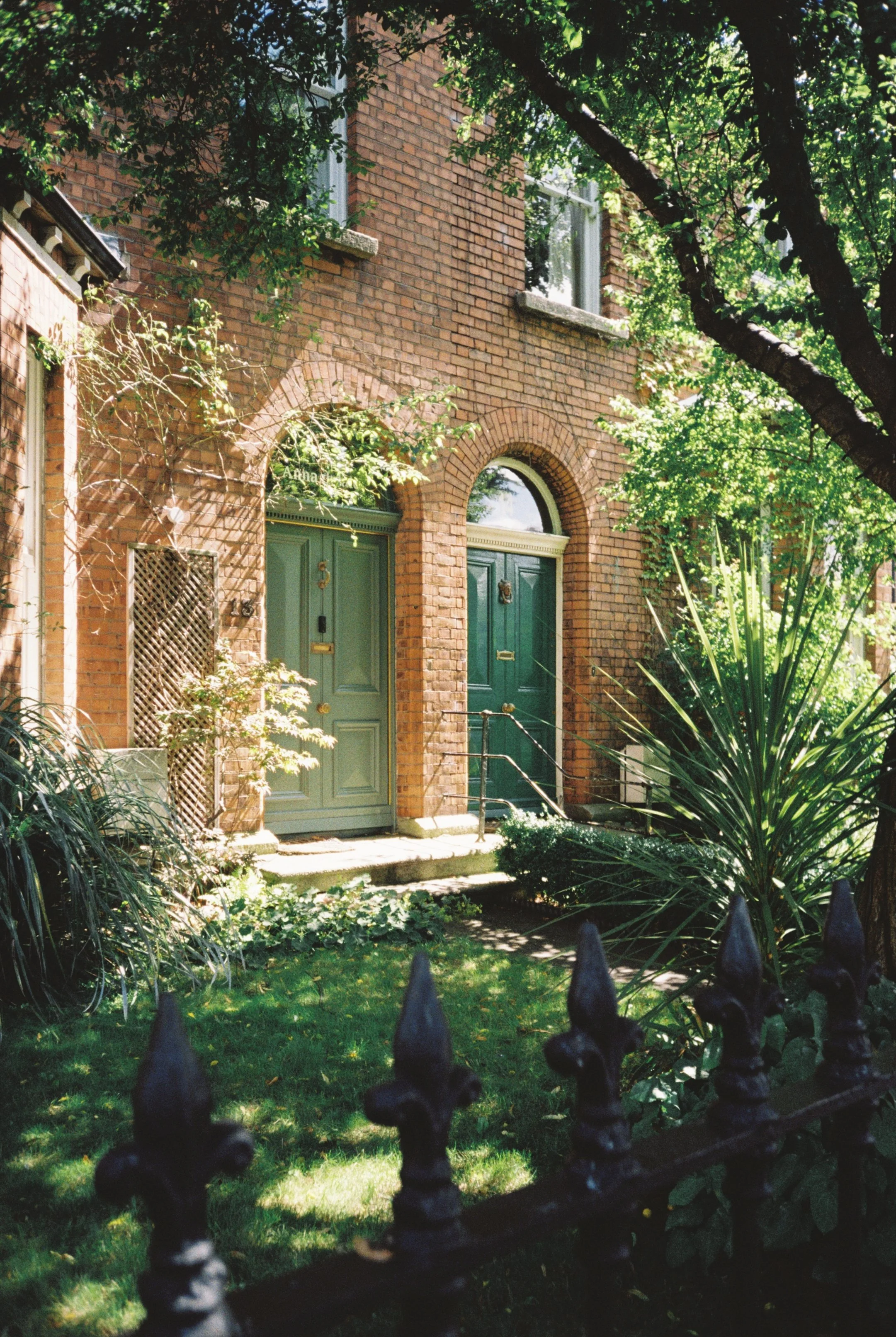 A brick townhouse with two green doors, surrounded by lush greenery and trees, with a black wrought iron fence in the foreground.