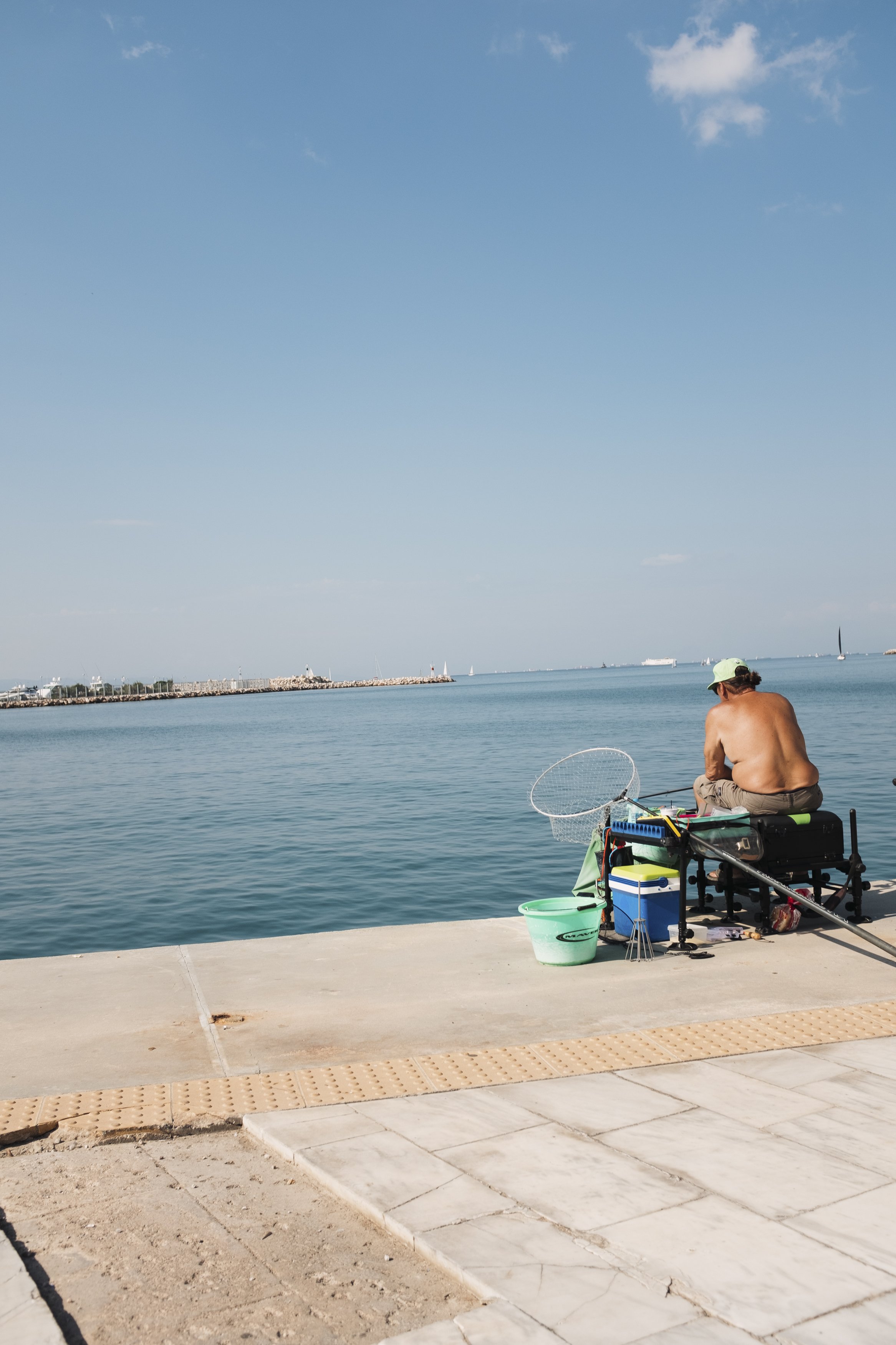 A man fishing on a concrete dock by the water, with fishing equipment and a cooler nearby, under a blue sky.