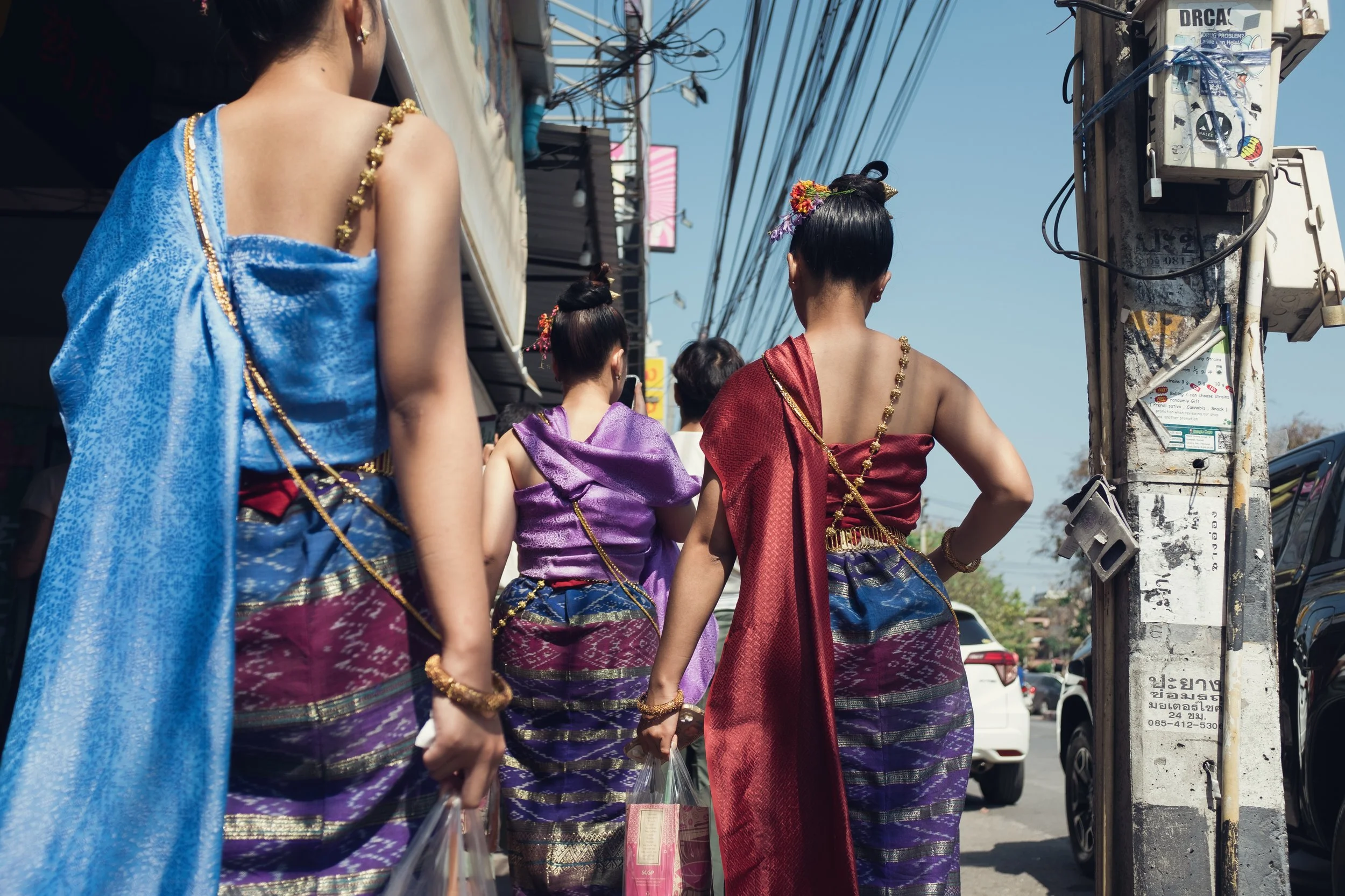 Four women dressed in traditional Thai attire walking on a street, wearing colorful silk clothing and gold jewelry, with a utility pole and power lines overhead.