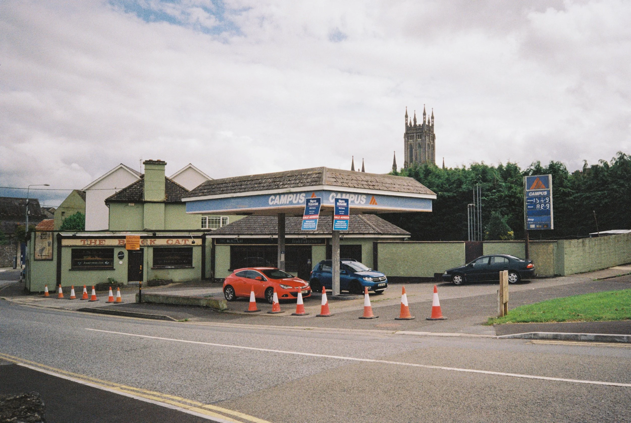 A small convenience store with a blue Canopus car wash canopy in front. There are three cars parked in the lot, with cones blocking part of the space. A church tower is visible in the background partially obscured by trees.
