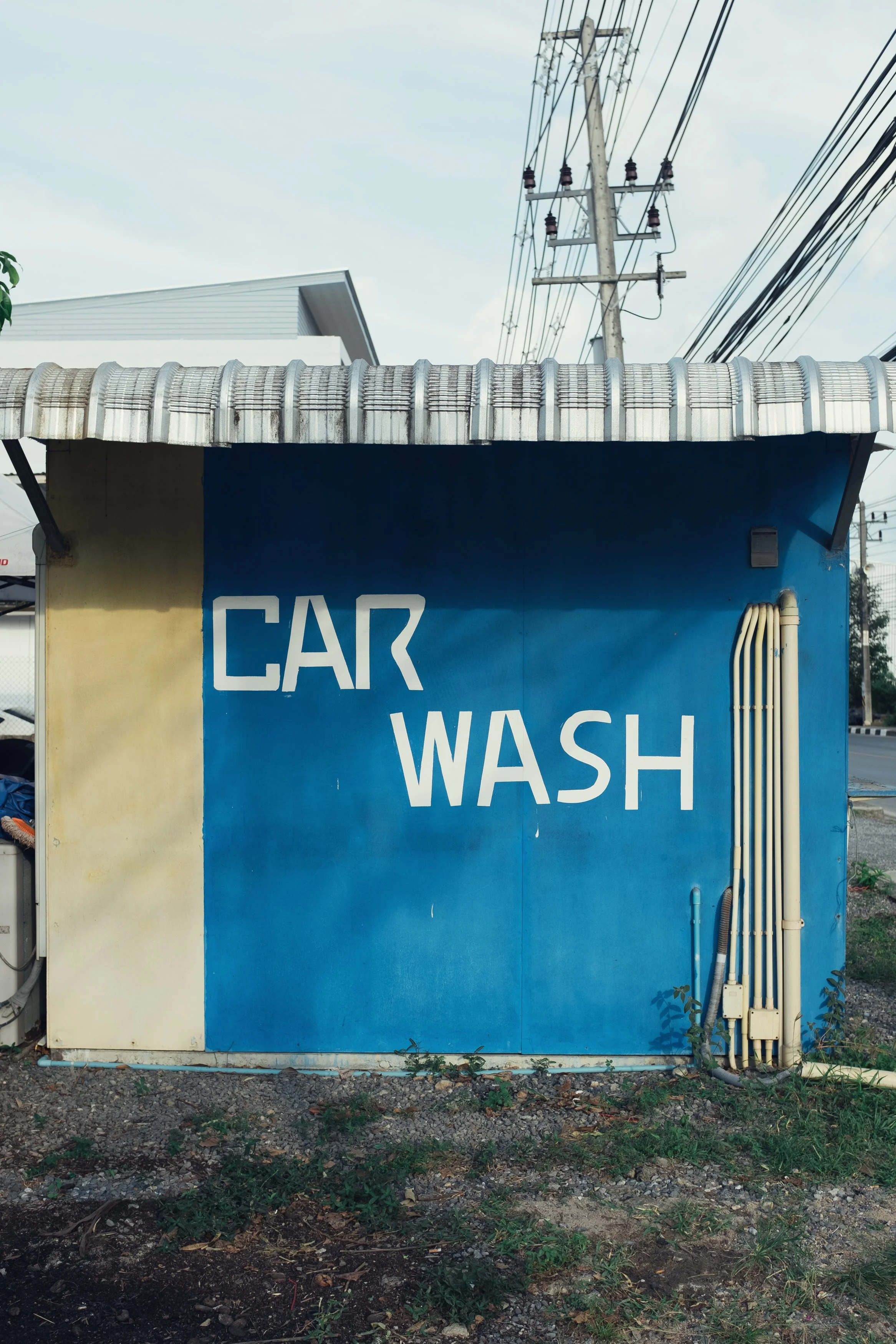A small building with a blue wall and a yellow section, with the words "CAR WASH" painted on it in white. The building has exposed pipes on the right side and a corrugated metal roof.