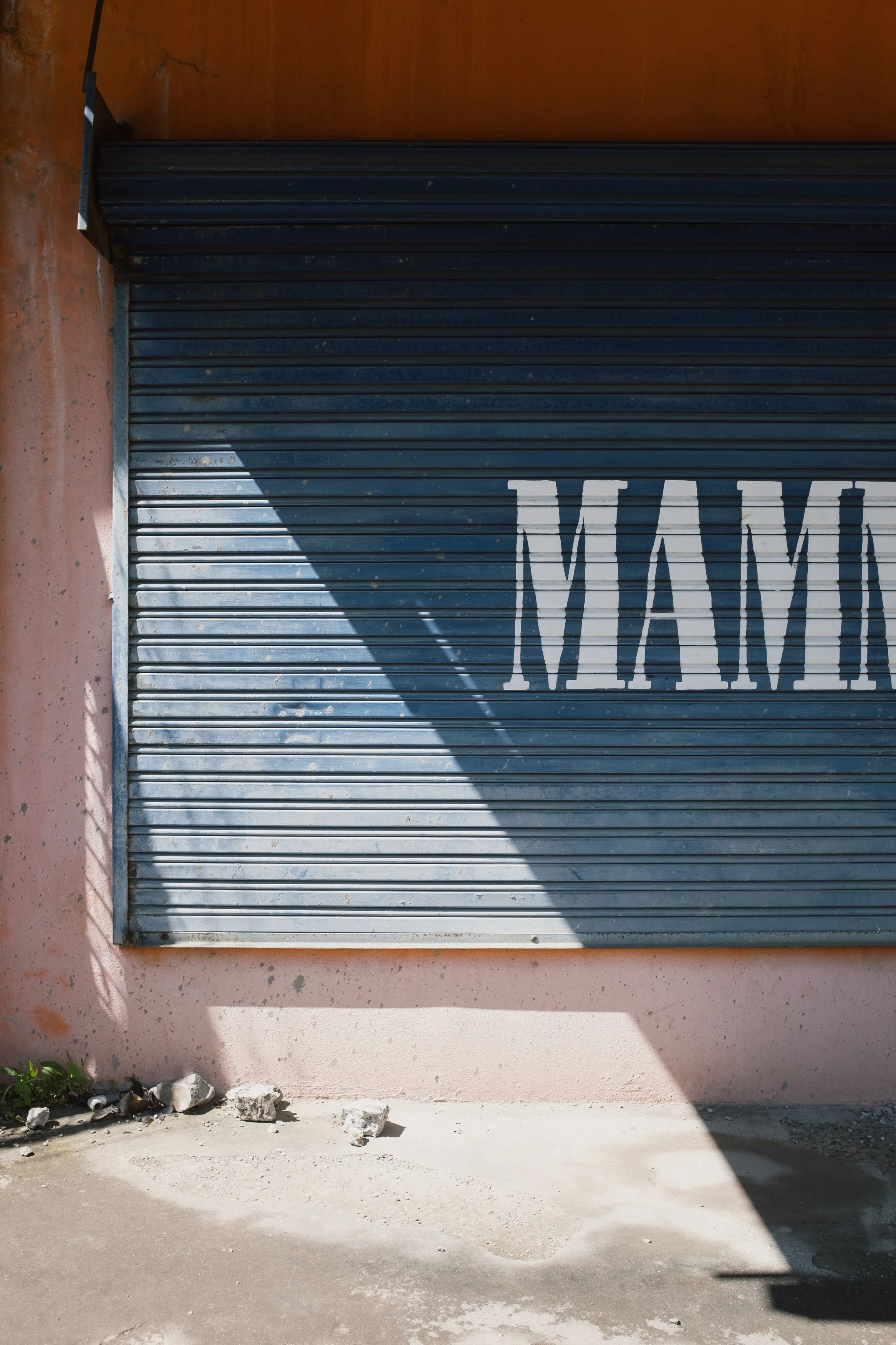 Close-up of a blue rolling shutter door with the word 'MAM' painted on it, casting a diagonal shadow and surrounded by a pink wall with some debris and rocks on the ground.