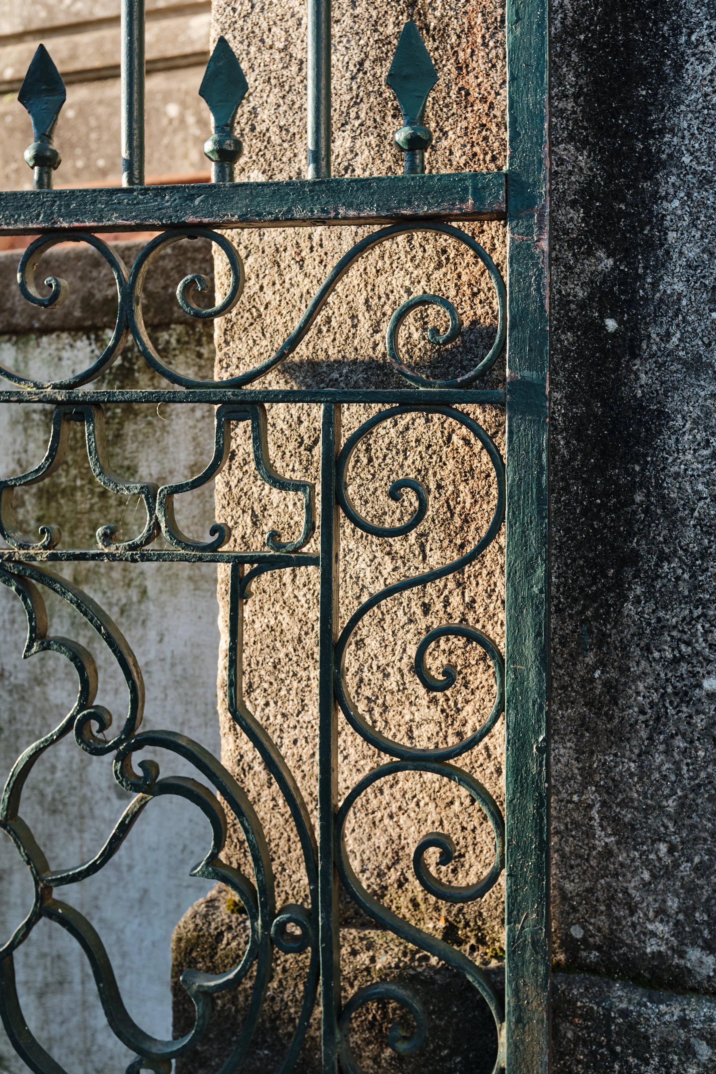 Close-up of an ornate, black wrought iron fence with decorative swirls and finials, mounted on a rough stone wall.