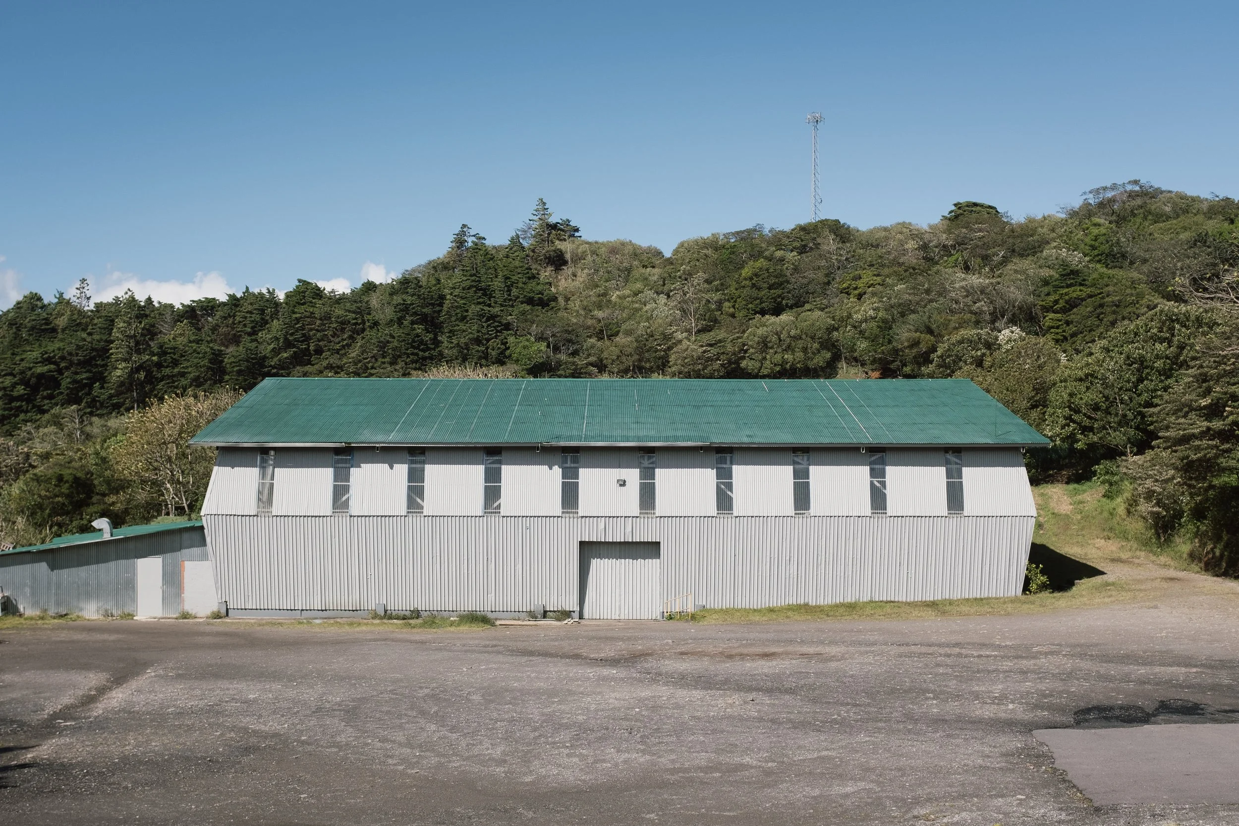 A large white industrial building with a green metal roof, situated in front of a hillside covered with trees under a clear blue sky.