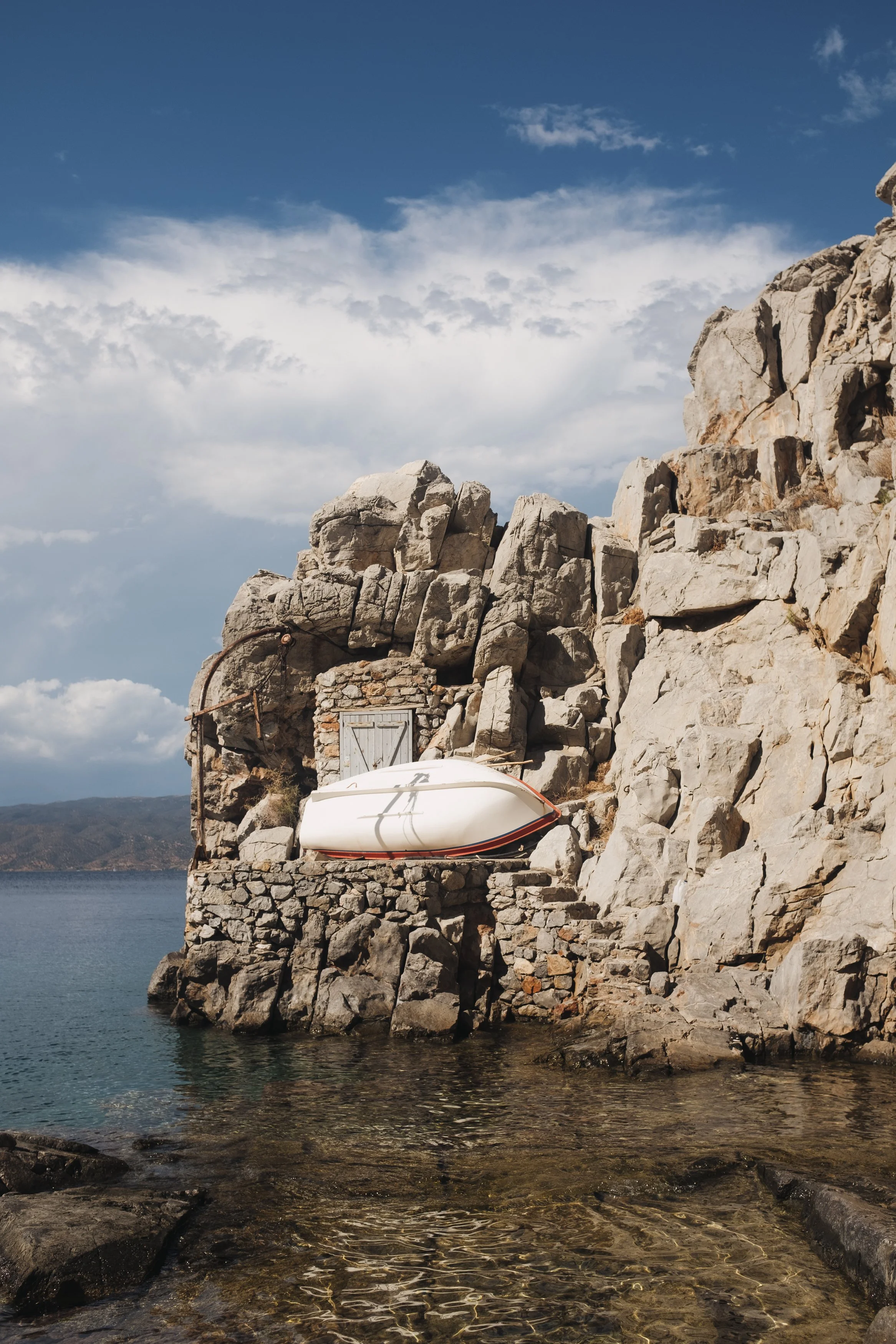 A small boat resting on a stone platform built into a rocky shoreline with a door behind it, overlooking a body of water under a partly cloudy sky.
