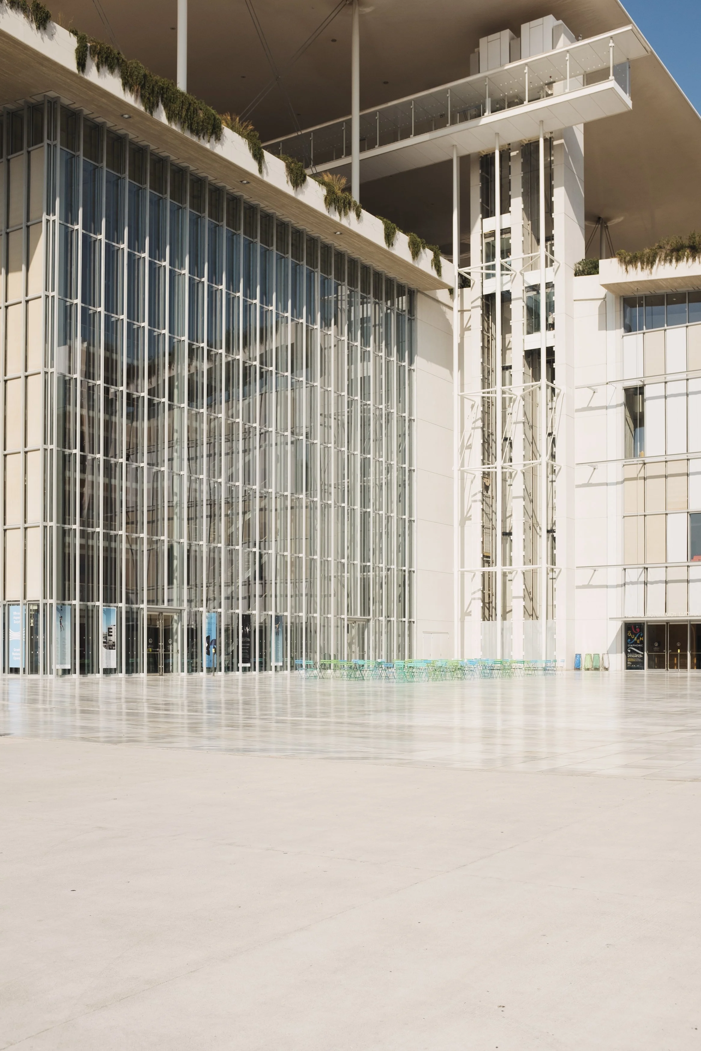Modern building with large glass windows, white structural elements, and green chairs outside, under a clear blue sky.