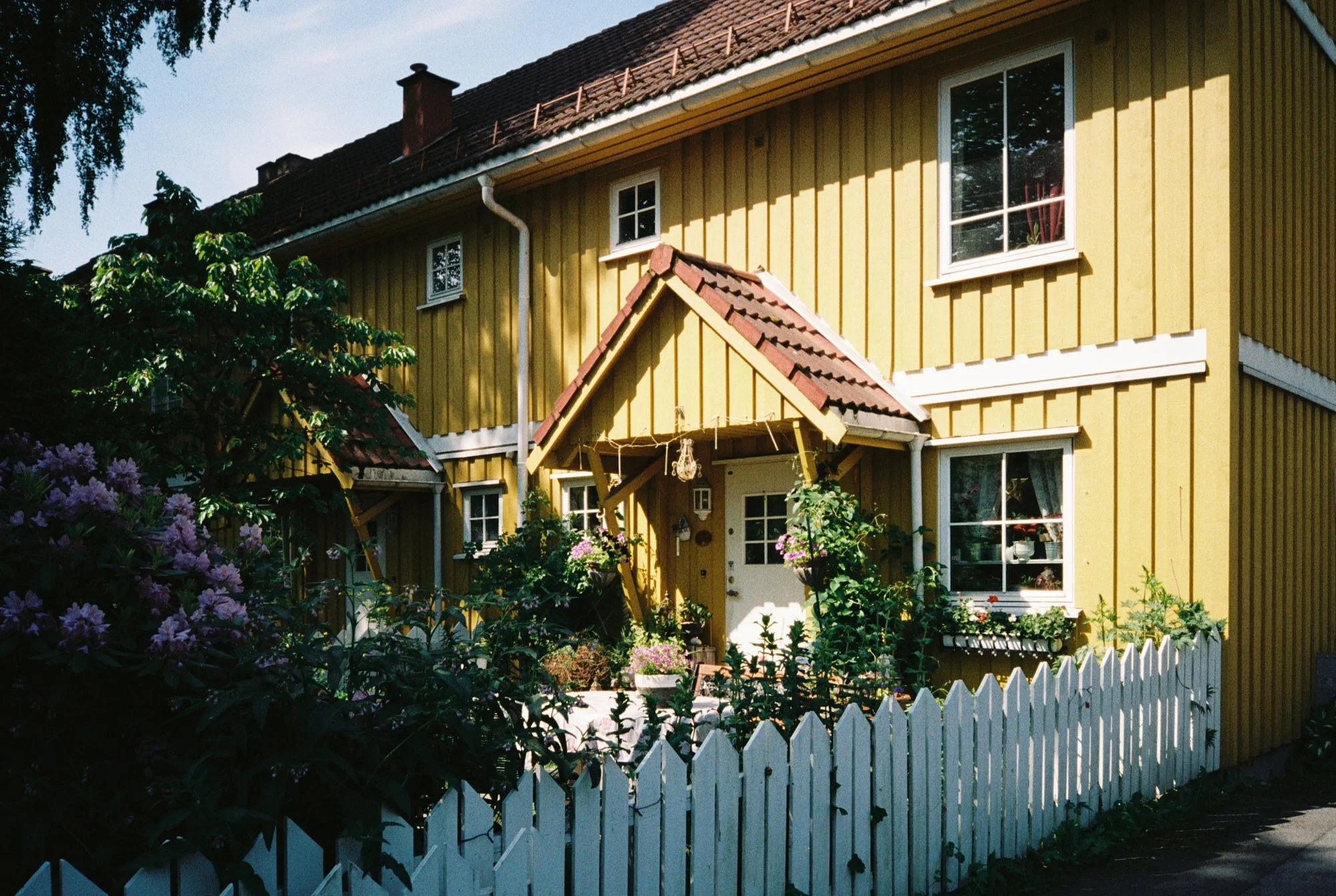 A yellow wooden house with white trim, a small front porch with a sloped roof, surrounded by a white picket fence and garden with blooming flowers.