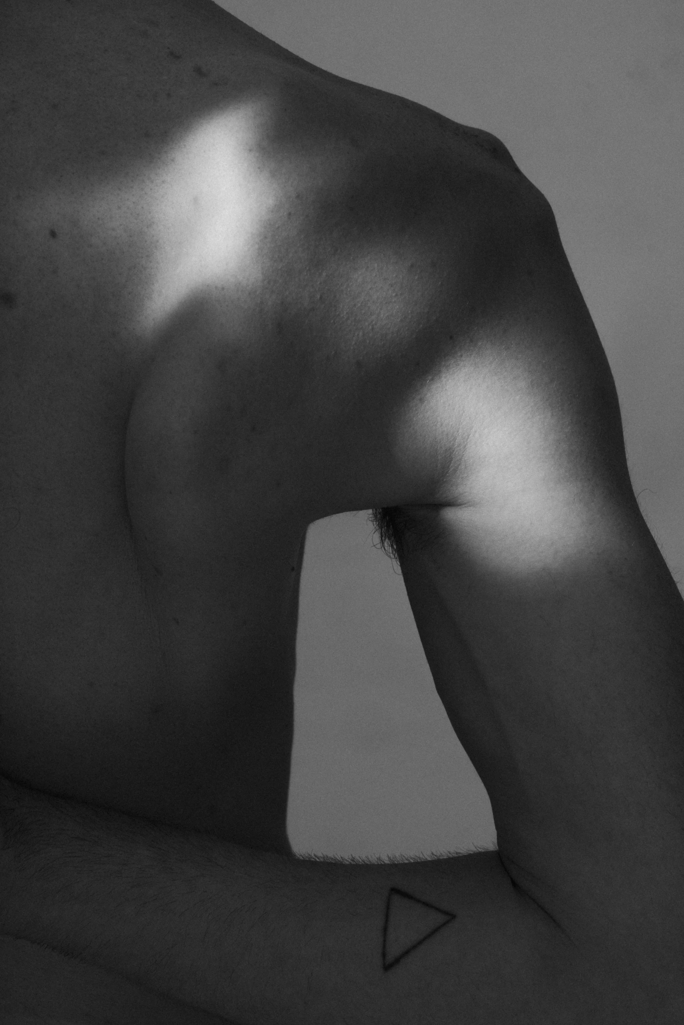 Close-up of a person’s bare shoulder, upper arm, and part of the chest in black and white, with a small triangle tattoo on the inner forearm.