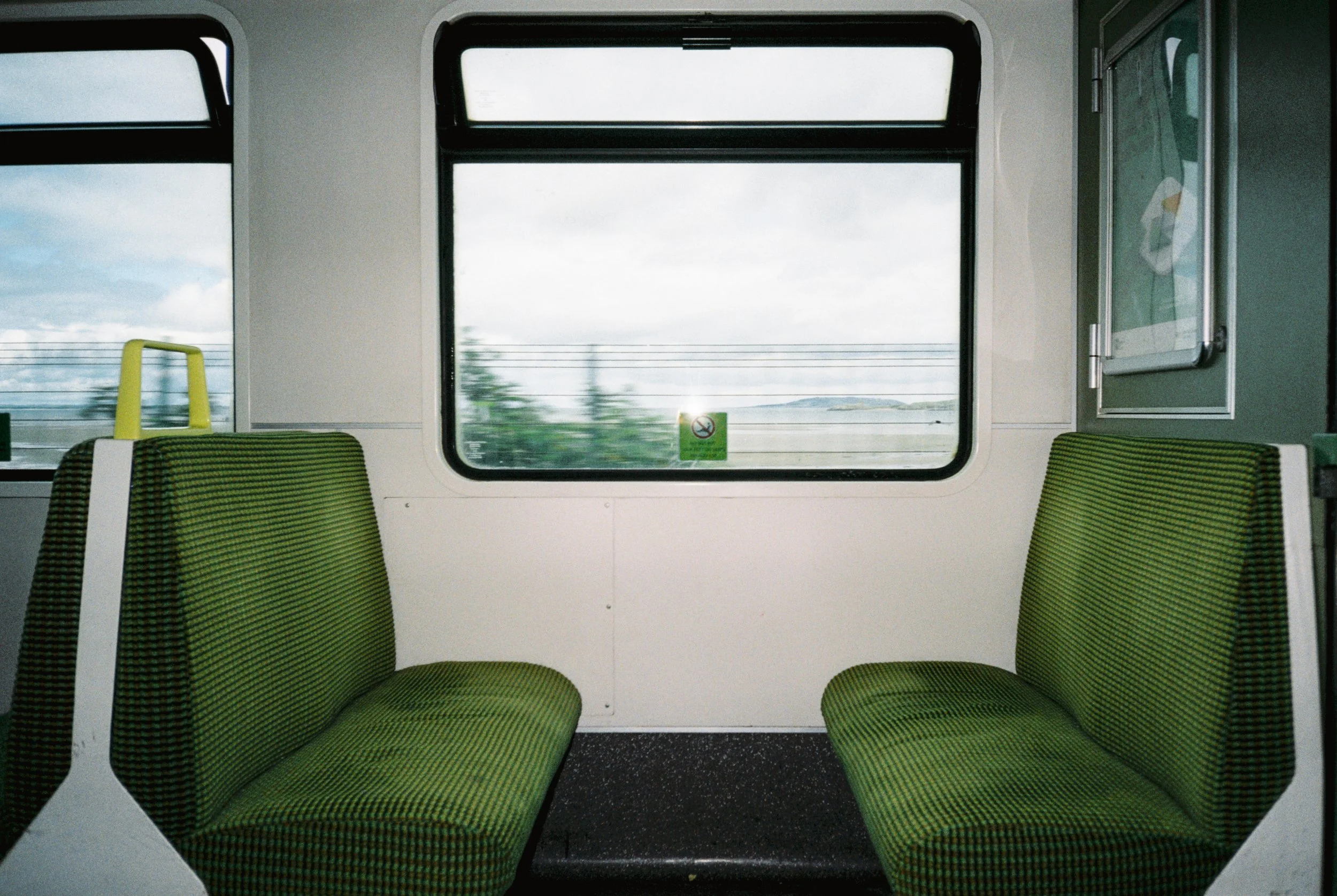 Inside a train car with two green upholstered seats facing each other, large window in the background showing a blurred landscape, and a small window to the right with a metallic frame.