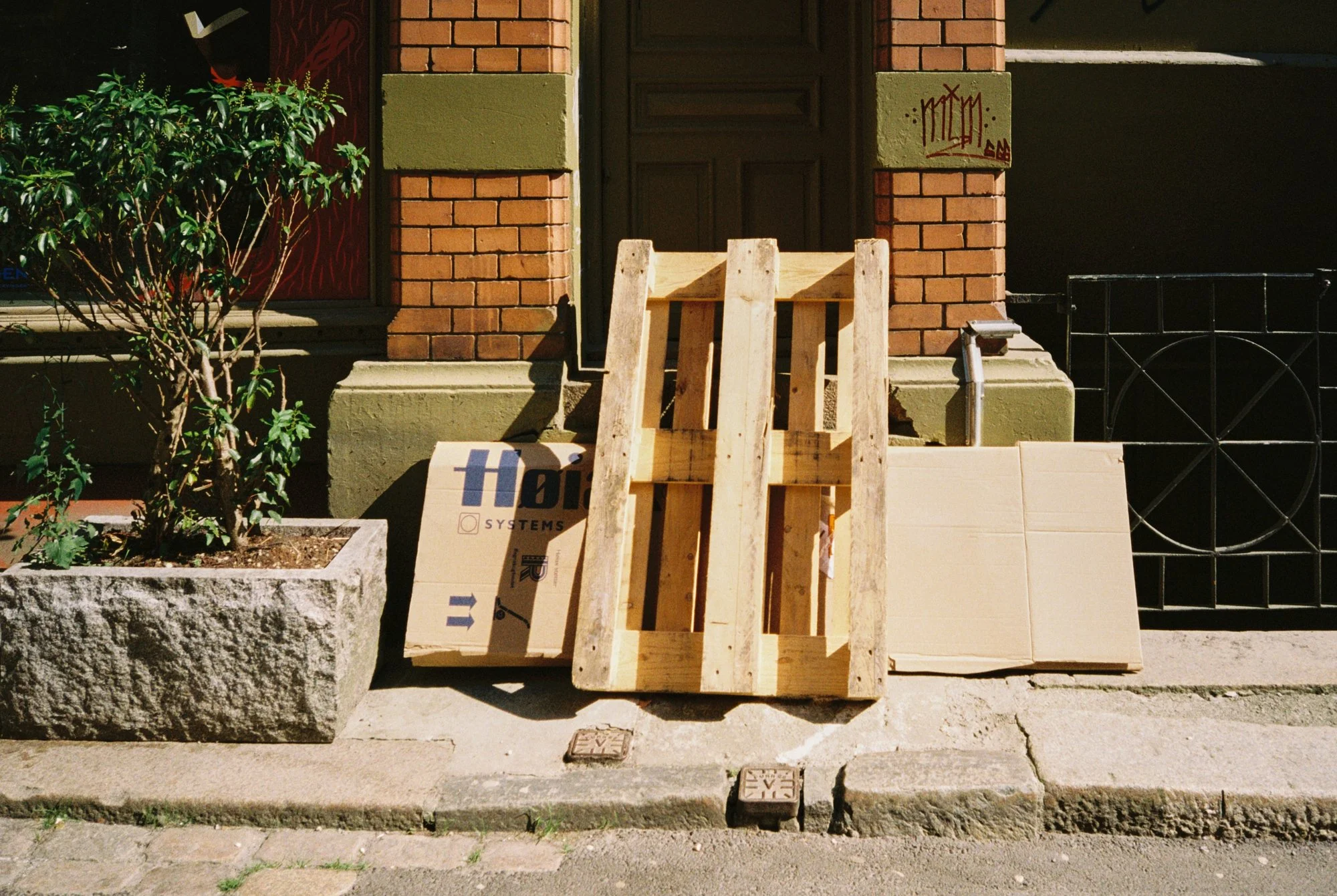 Street scene showing a wooden pallet, cardboard boxes, and a piece of plywood leaning against the sidewalk in front of a brick building with a dark door and green trim, and a small shrub in a concrete planter.