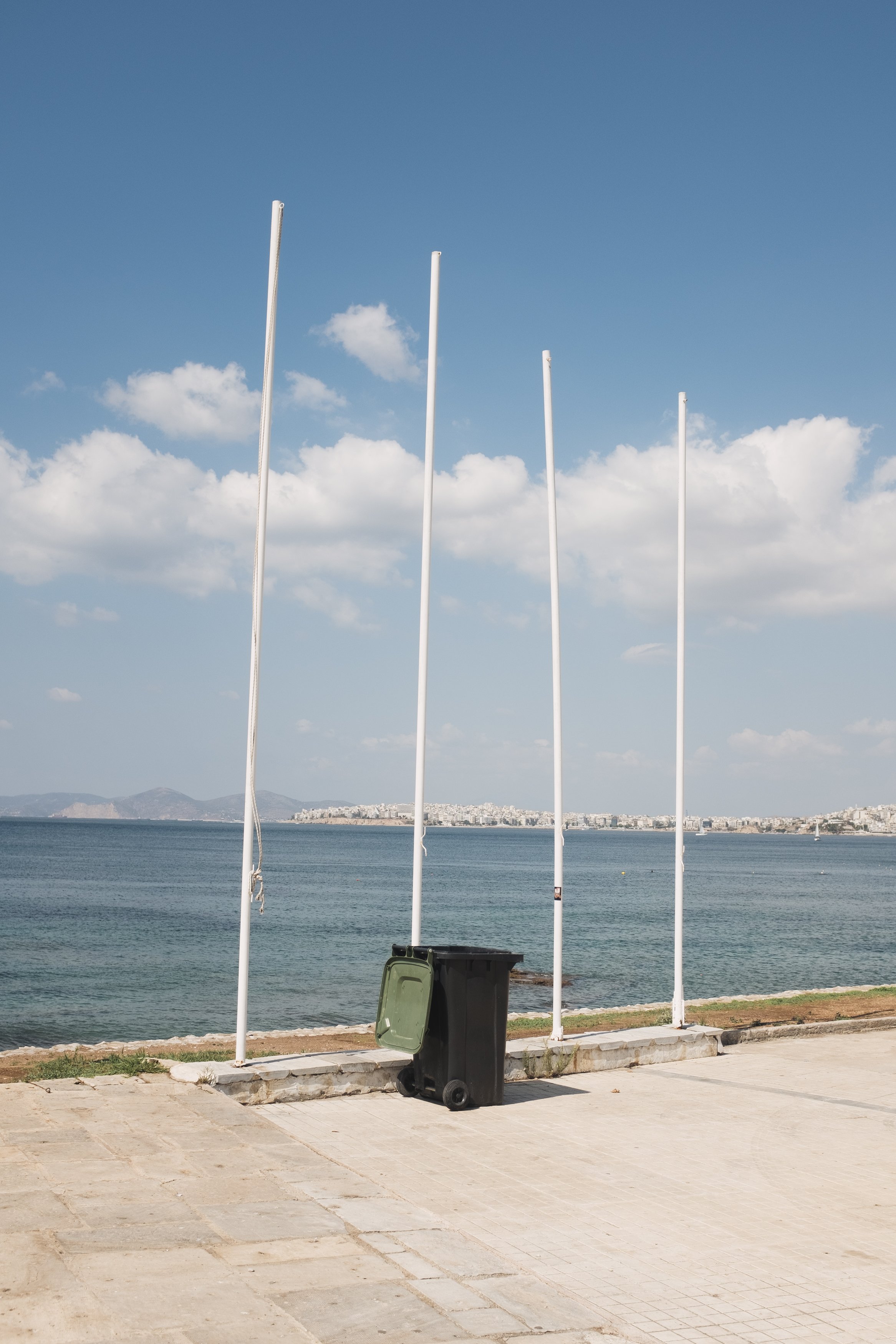 Empty flagpoles near water with a black trash bin in the foreground and a distant city across the bay under a partly cloudy sky.
