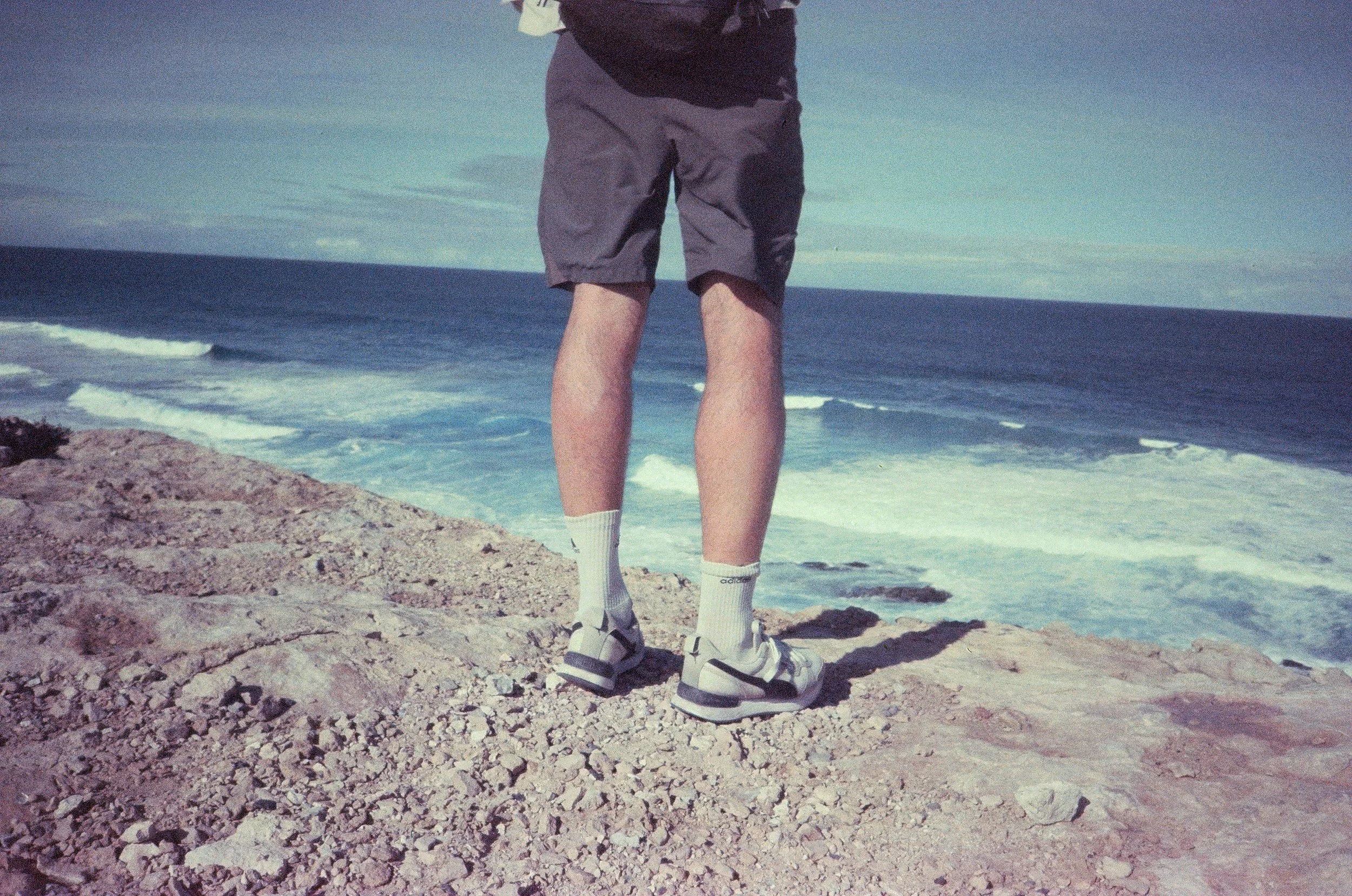 Person standing on rocky cliff overlooking the ocean, wearing gray shorts, white socks, and white sneakers, with the ocean and sky in the background.