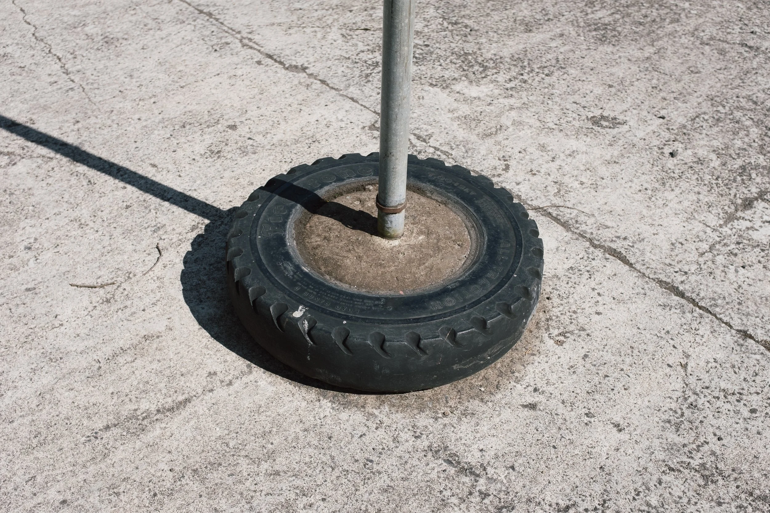 Close-up of a metal pole anchored into a rubber tire base on a concrete surface, with a shadow cast to the left.