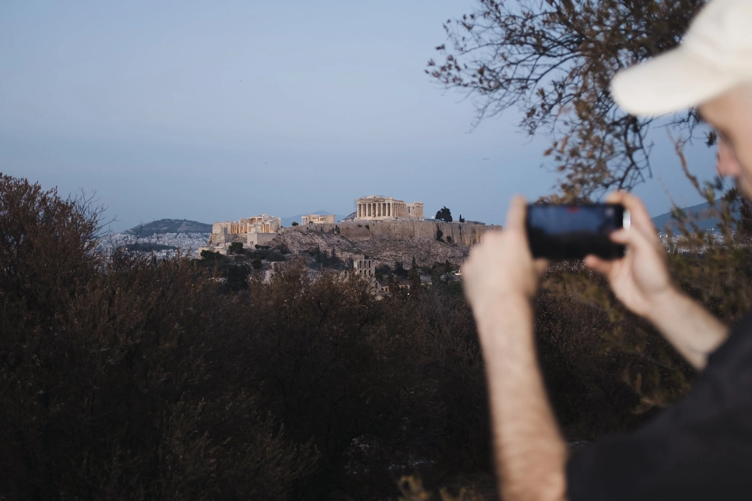 A person taking a photo of the Parthenon on the Acropolis in Athens, Greece, during dusk.