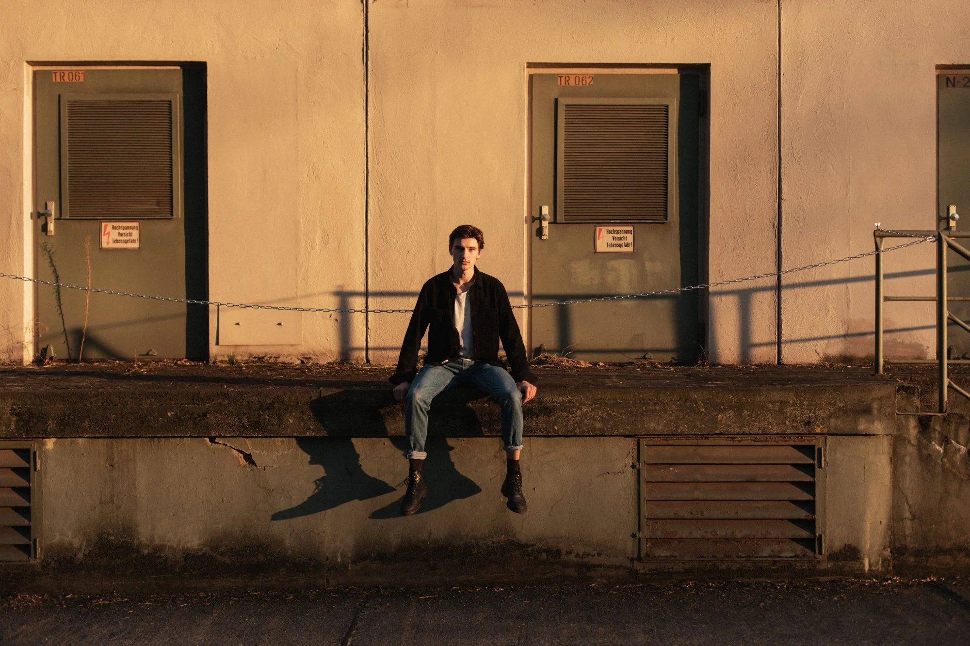 A young man sitting on a ledge in front of a beige building with three gray doors at sunset.