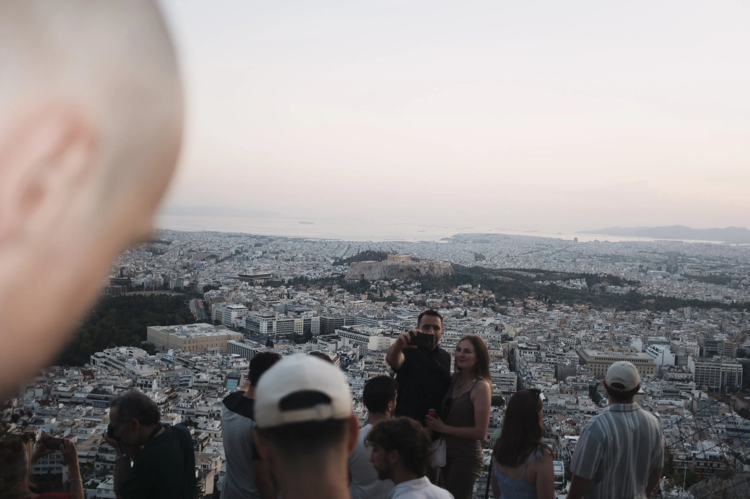 People taking photos and enjoying the view of a cityscape from an elevated vantage point, with a blurred foreground and a historical site in the distance.