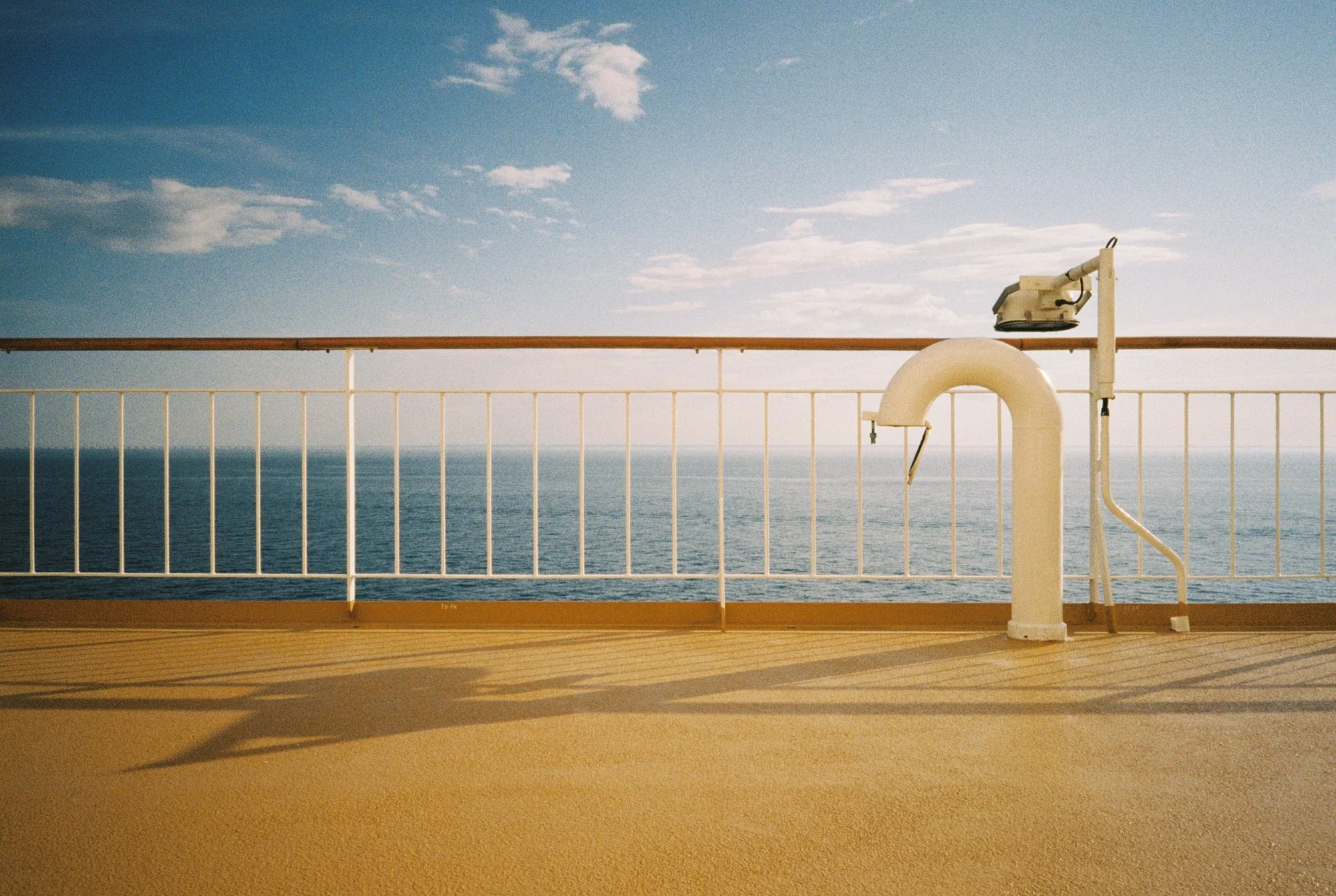 View of the ocean from a ship deck with a white railing, a coin-operated binocular viewer, and a shadow cast on the tan deck under a partly cloudy sky.