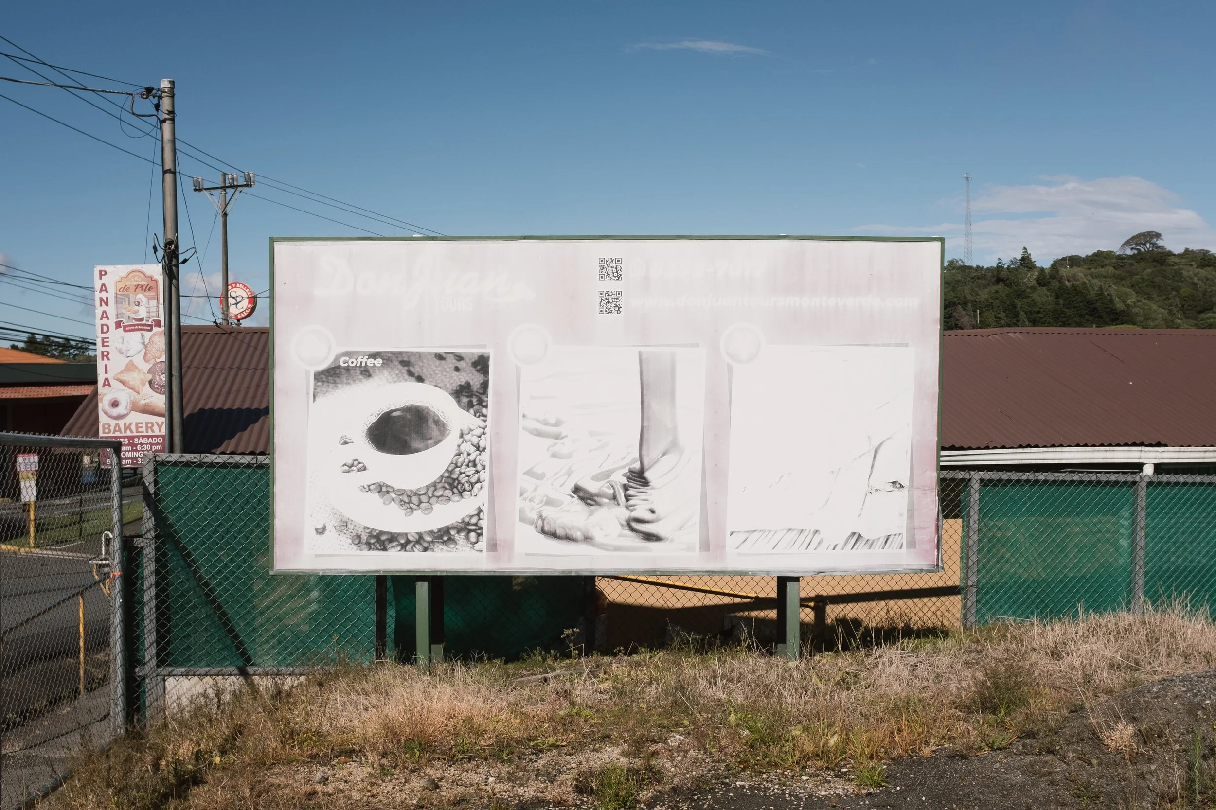 A billboard with faded and unclear images and text stands beside a fenced area with a bakery sign on a pole, under clear blue sky and nearby trees.