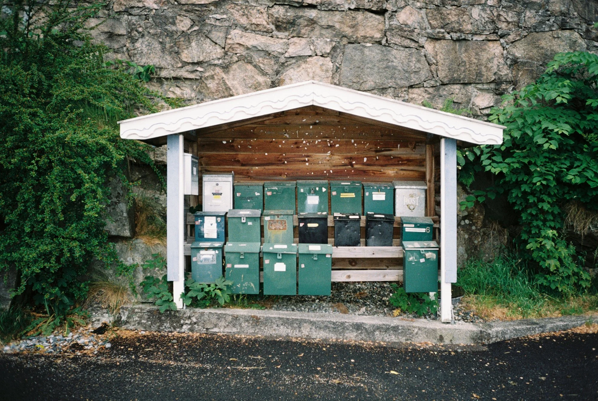 A small shelter with a white roof and wooden back wall, housing multiple green and black mailboxes, situated against a rock wall with green foliage on either side.