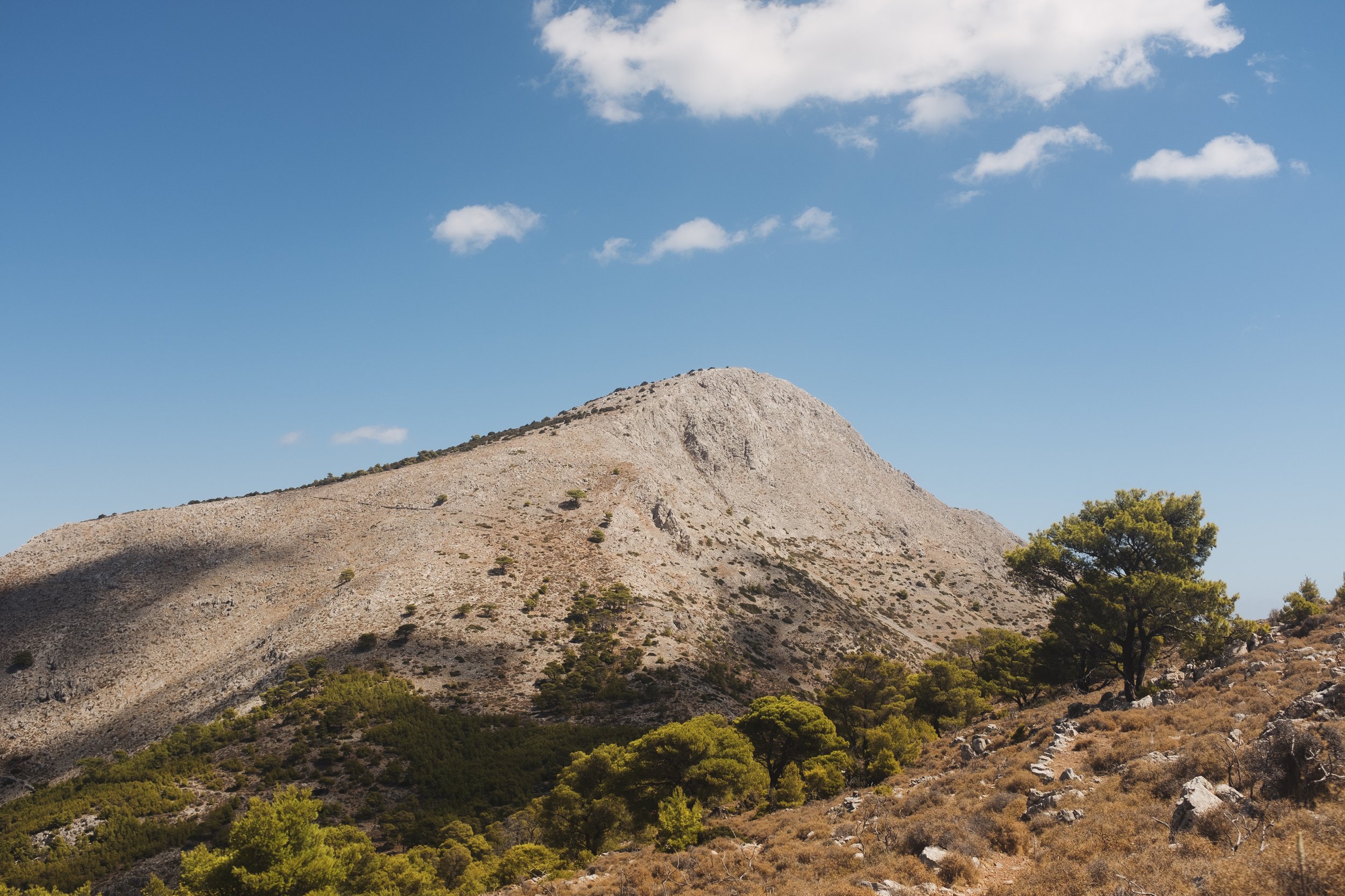 A mountain landscape with a large rocky hill, sparse vegetation, and a few green trees under a partly cloudy blue sky.