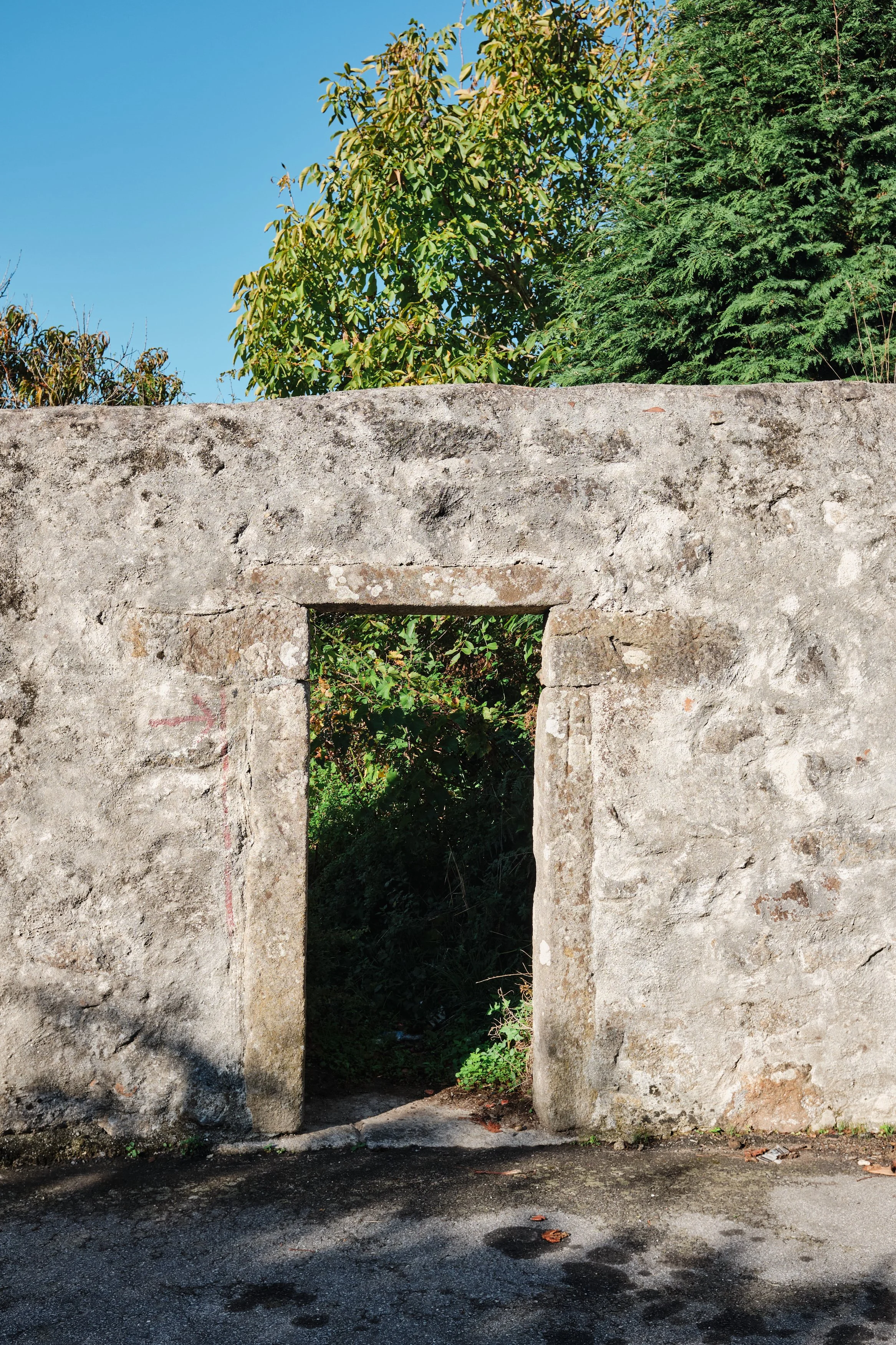 Old stone wall with a small rectangular opening, greenery visible through the opening, and trees in the background under a clear blue sky.