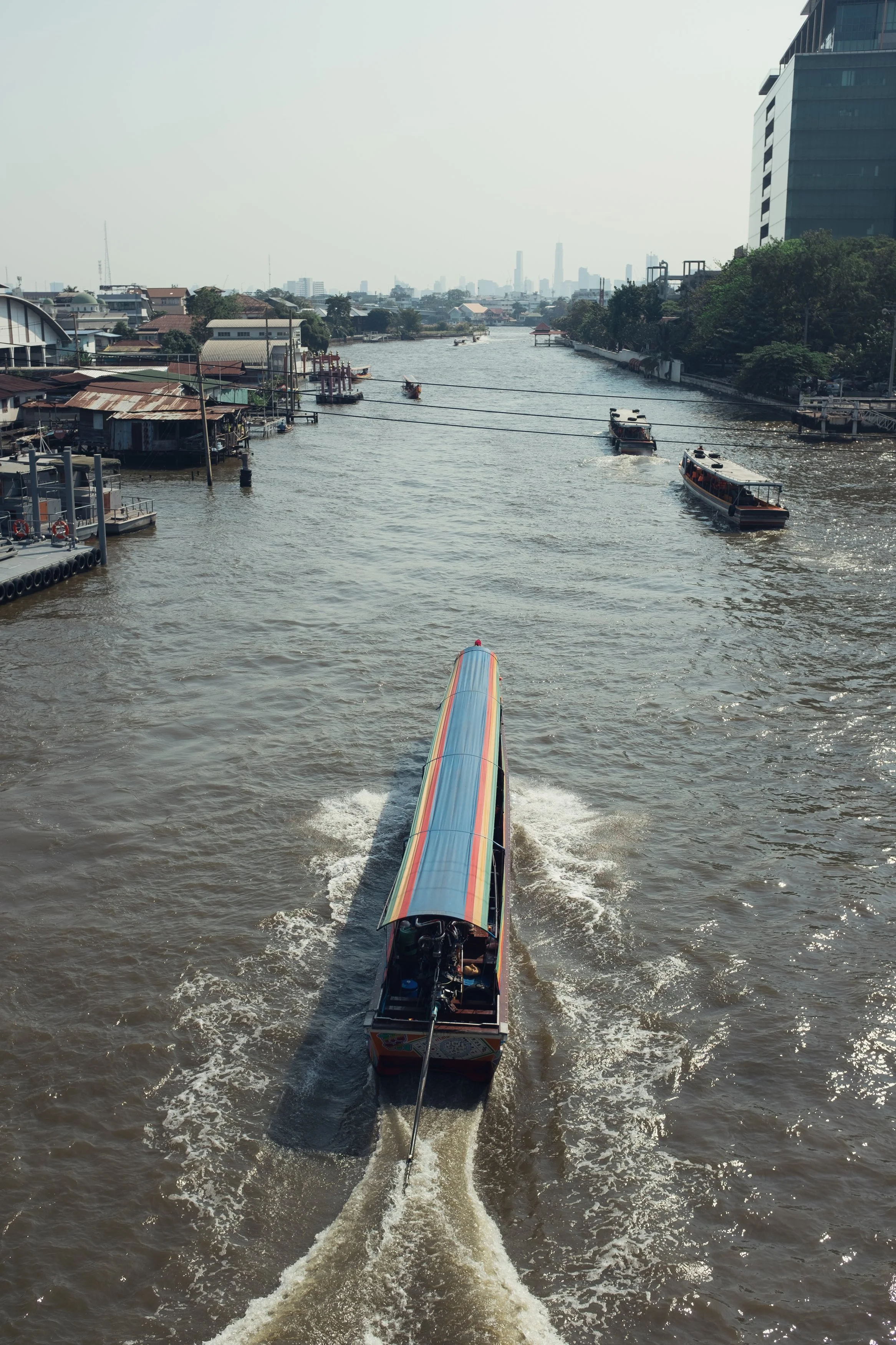A long boat with a colorful roof moving down a river in an urban area with boats and buildings on either side.