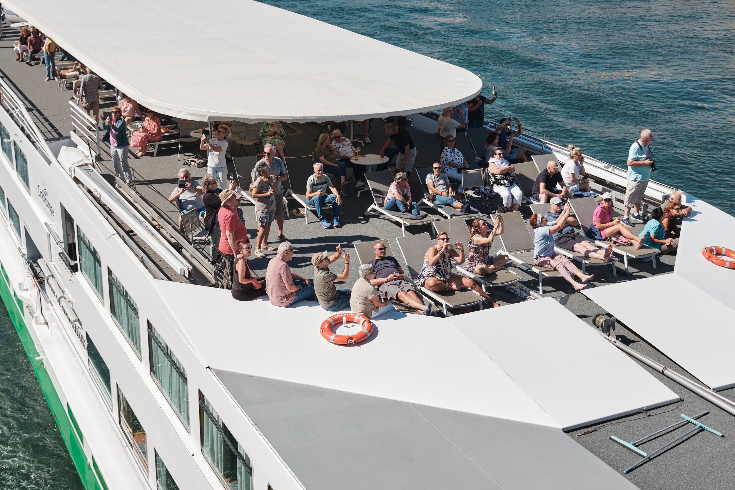 Crowd of people relaxing on the deck of a boat, with some sitting on lounge chairs, taking photos, and others standing or walking around, with water in the background.