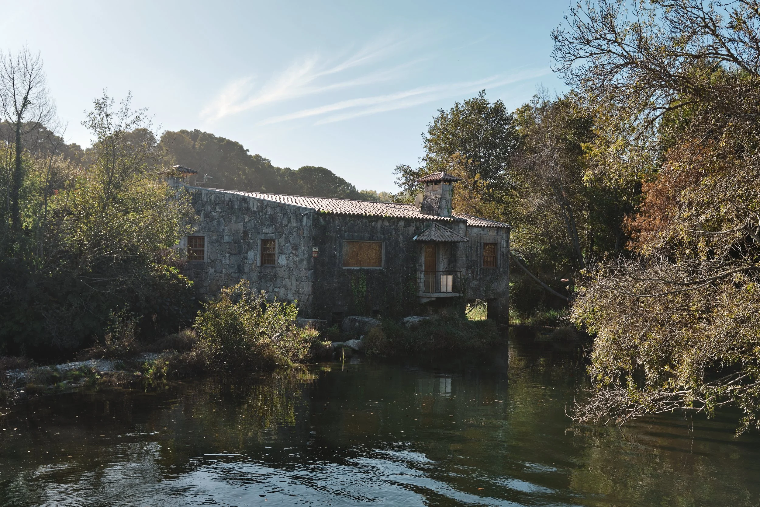 Stone house beside a river with trees and hills in the background under a blue sky.