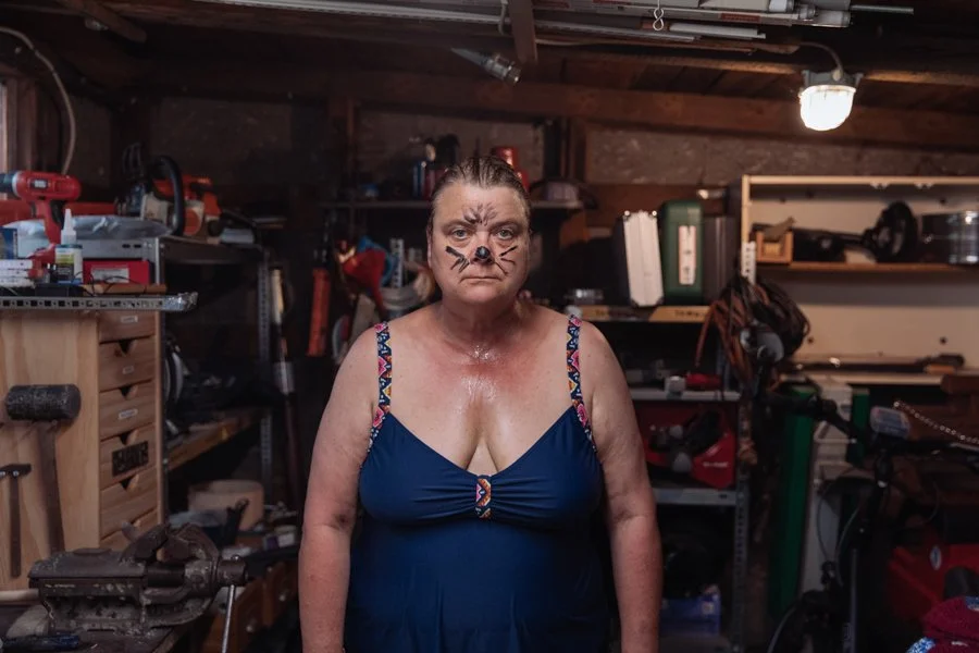A woman with face makeup resembling a cat, standing in a cluttered workshop or garage.