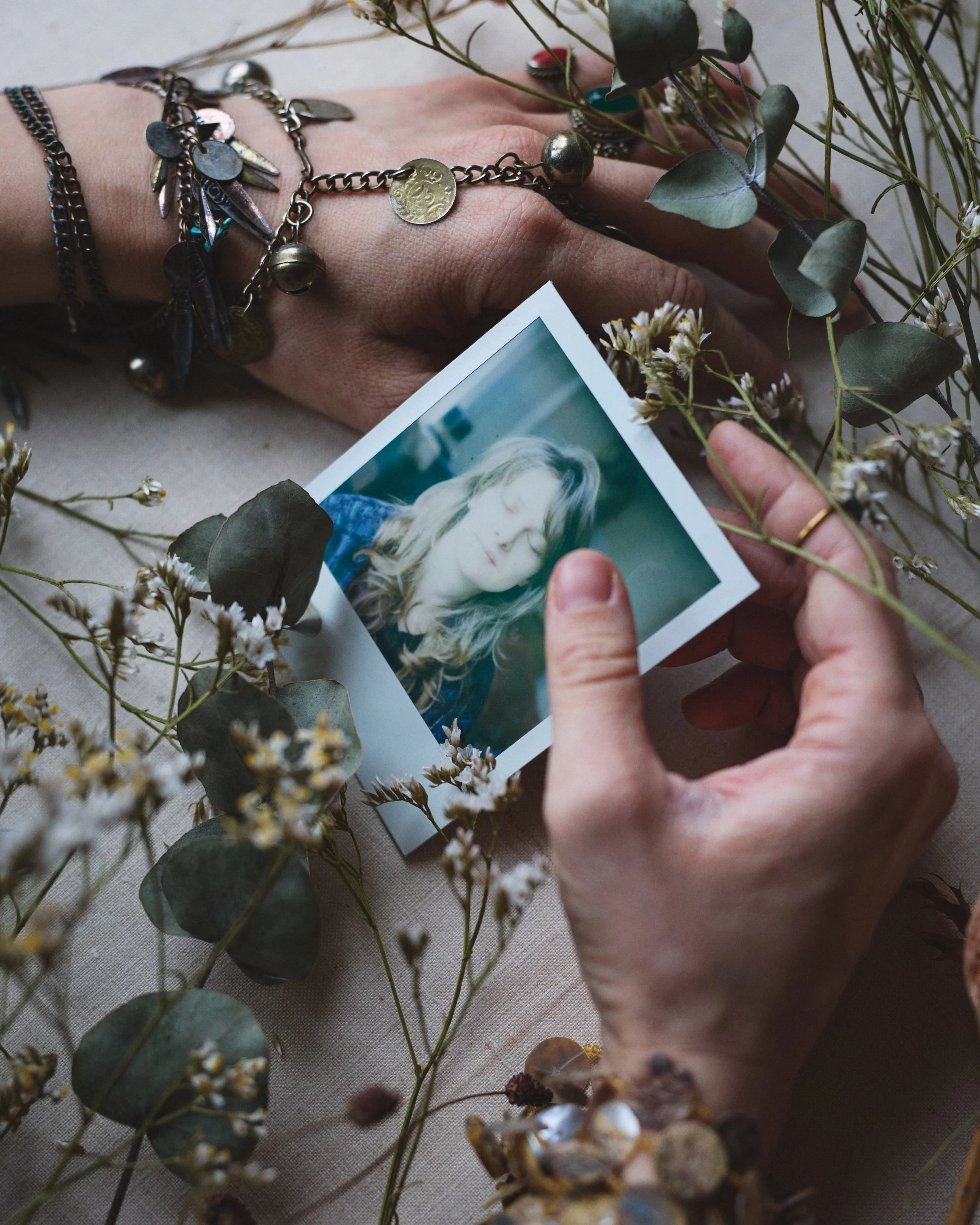 A person holding a photograph of a woman with long blonde hair amidst scattered greenery and small white flowers.