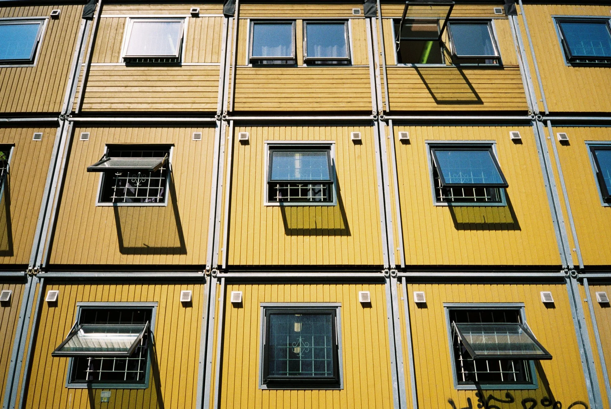 The image shows a modern apartment building facade with yellow wooden panels, multiple open windows with metal bars, and shadows cast by open window shutters.