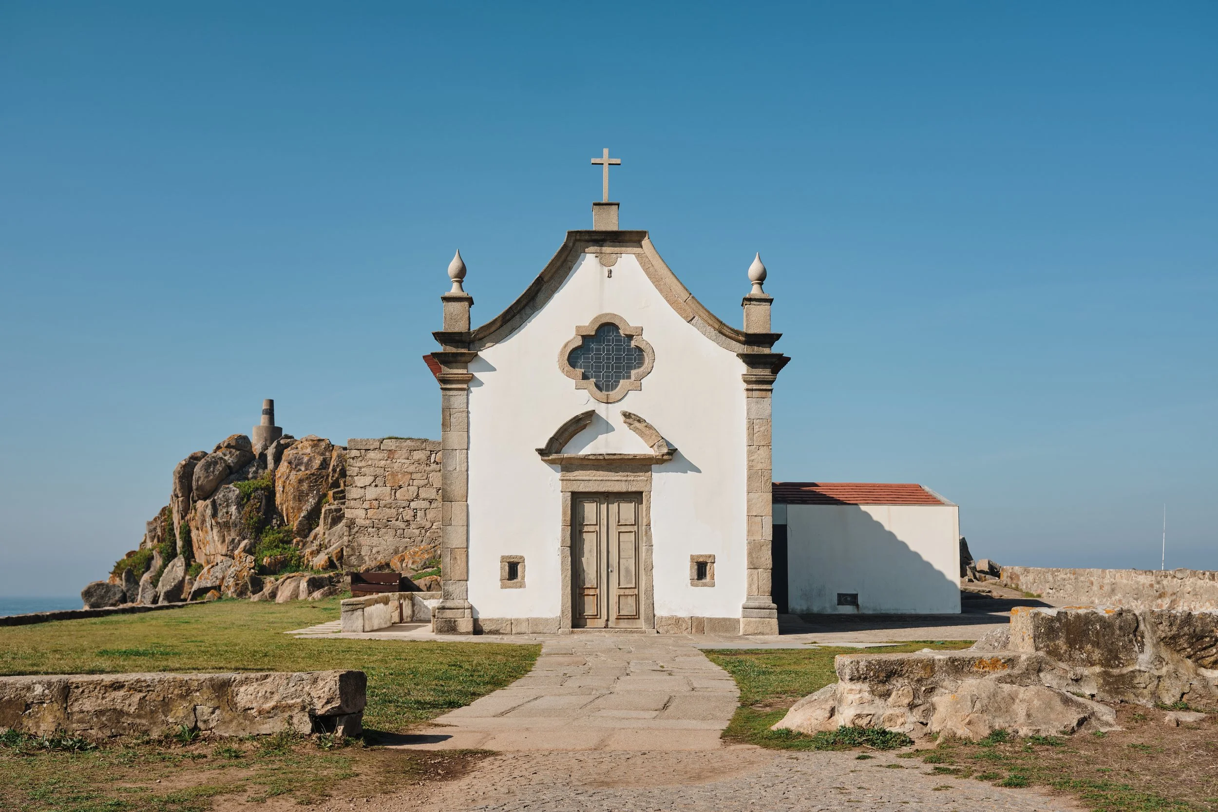 Small white chapel with a cross on top, stone frame around the door, and decorative window, situated outdoors on grassy terrain with large rocks and a clear blue sky.