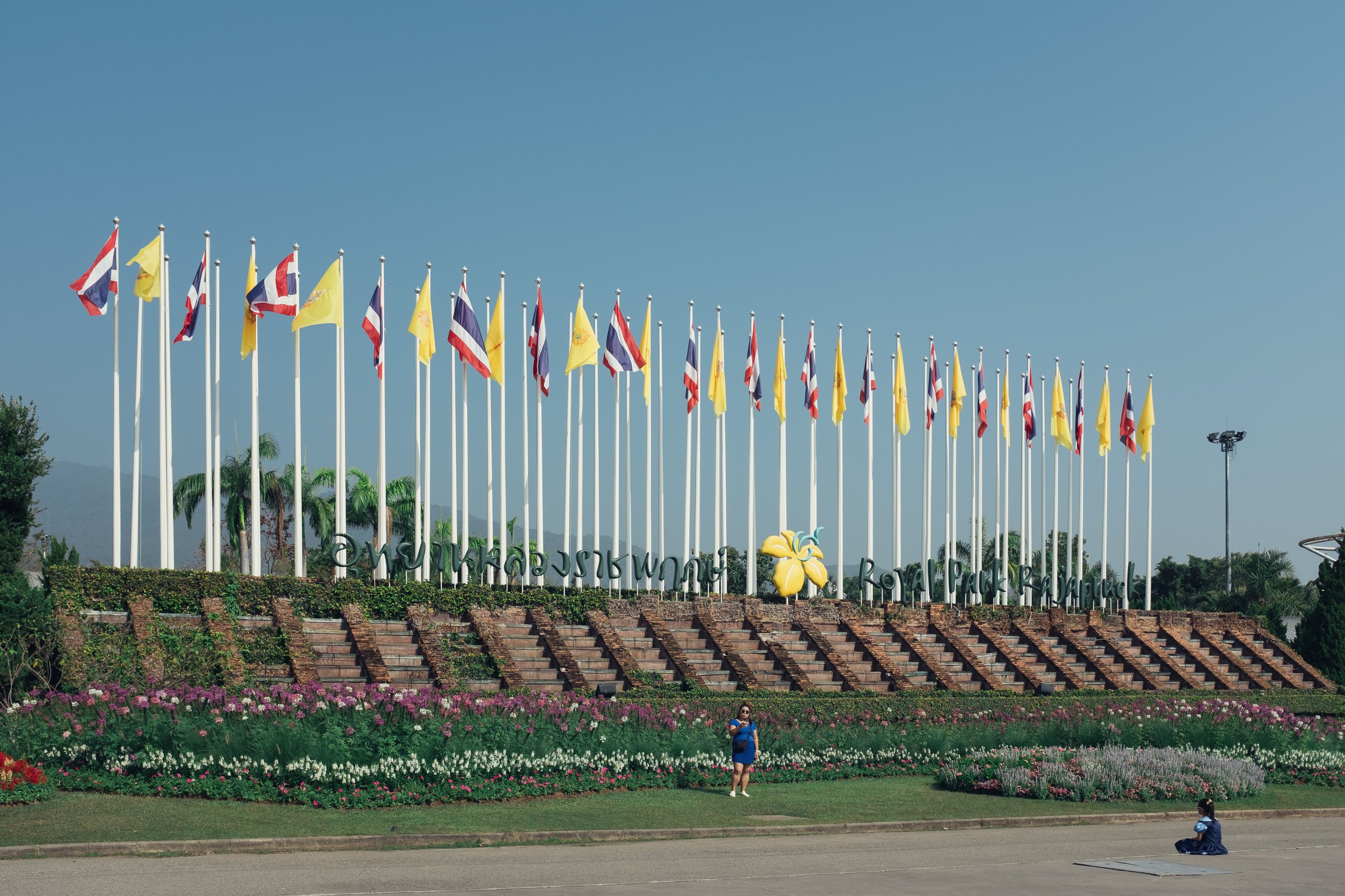 Flag display at Royal Park Rajapruek with colorful flowers and a person in blue dress taking a photo, and another person sitting on the ground in front.