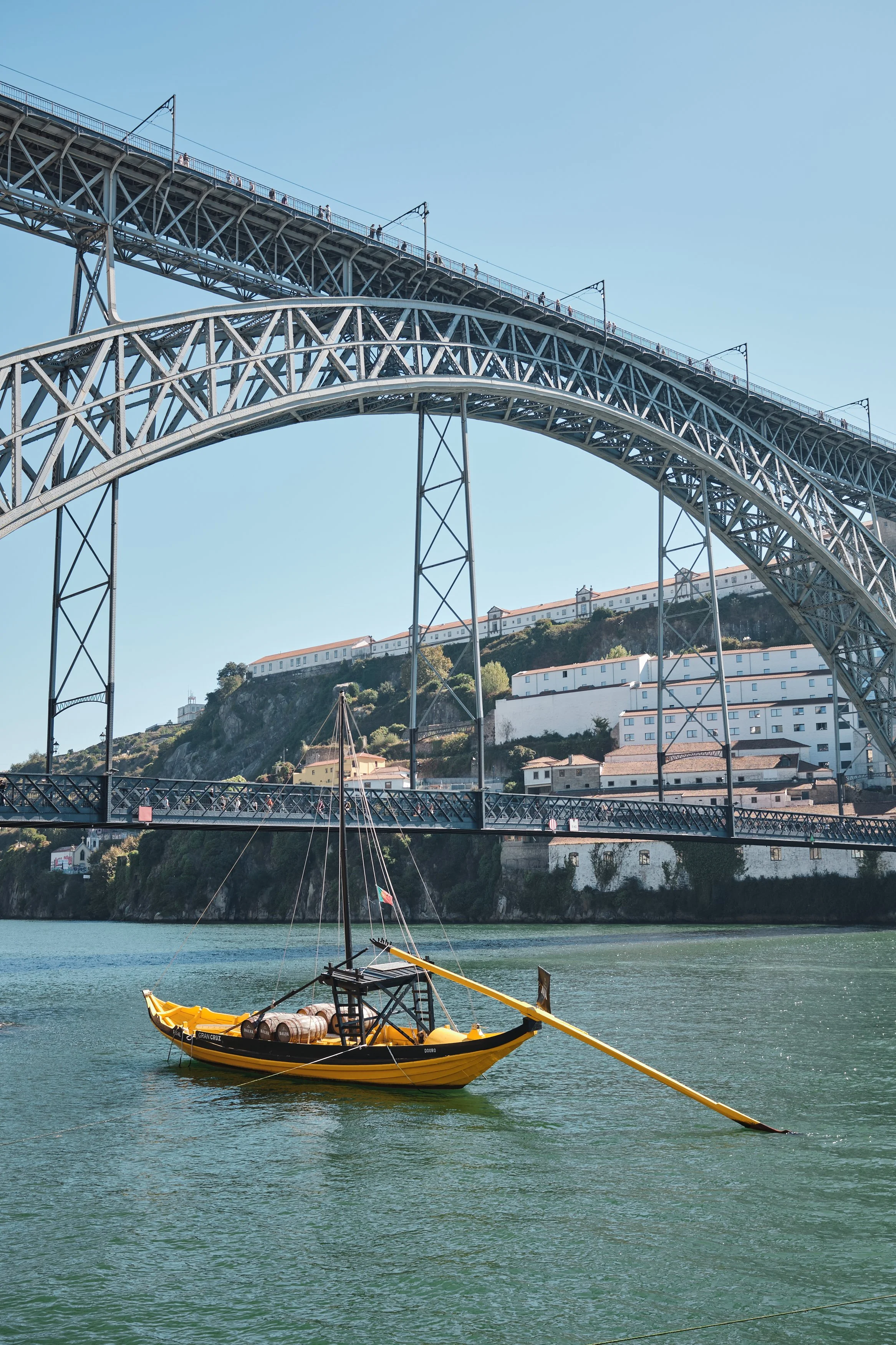 A yellow and black boat floating on water underneath a large steel arch bridge, with white buildings on a hillside in the background.