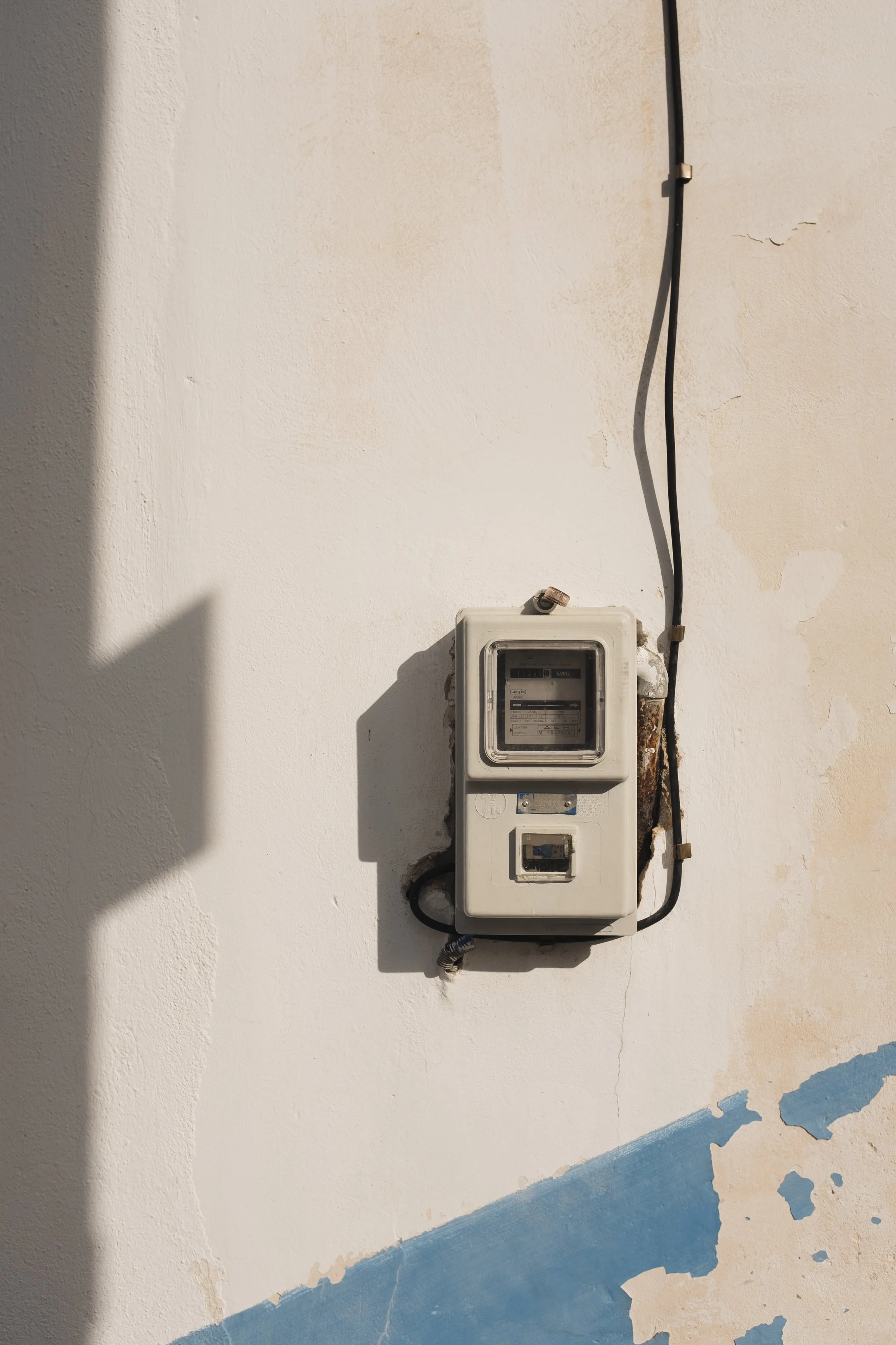 An outdoor wall with an old, weathered electric meter box, exposed wiring, and peeling paint near the bottom.