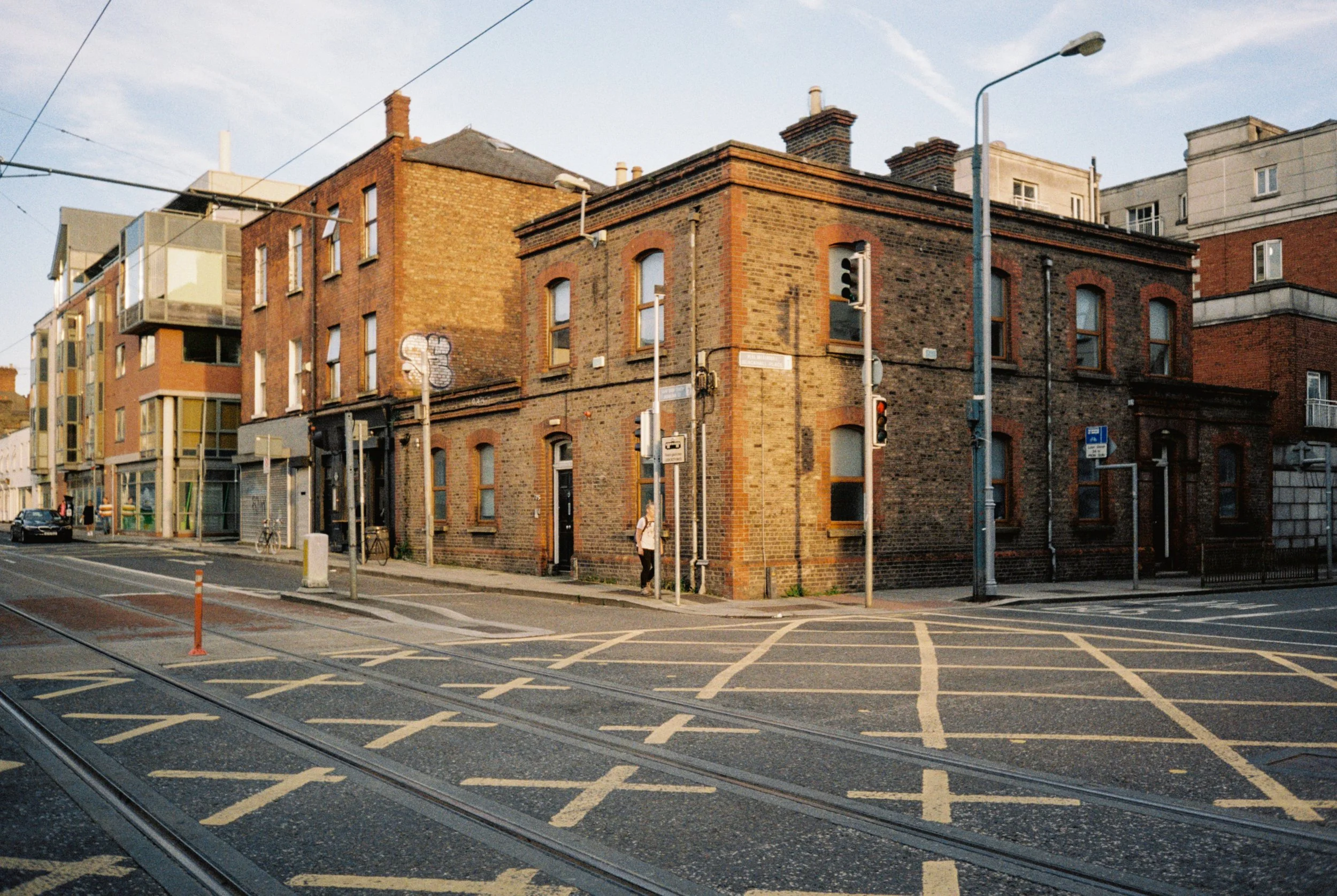 A city street scene with old brick buildings on the corner, tram tracks on the street, and several traffic lights and street signs, during sunset.