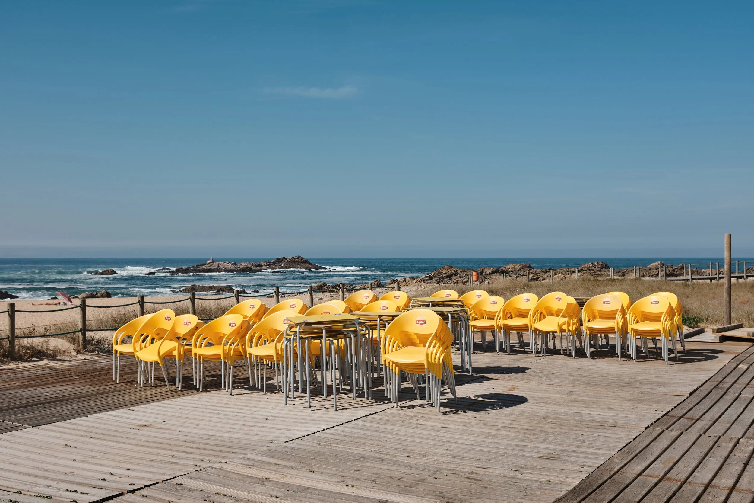 Stacked yellow chairs and tables on a wooden deck near the beach with rocks and ocean waves in the background under a blue sky.