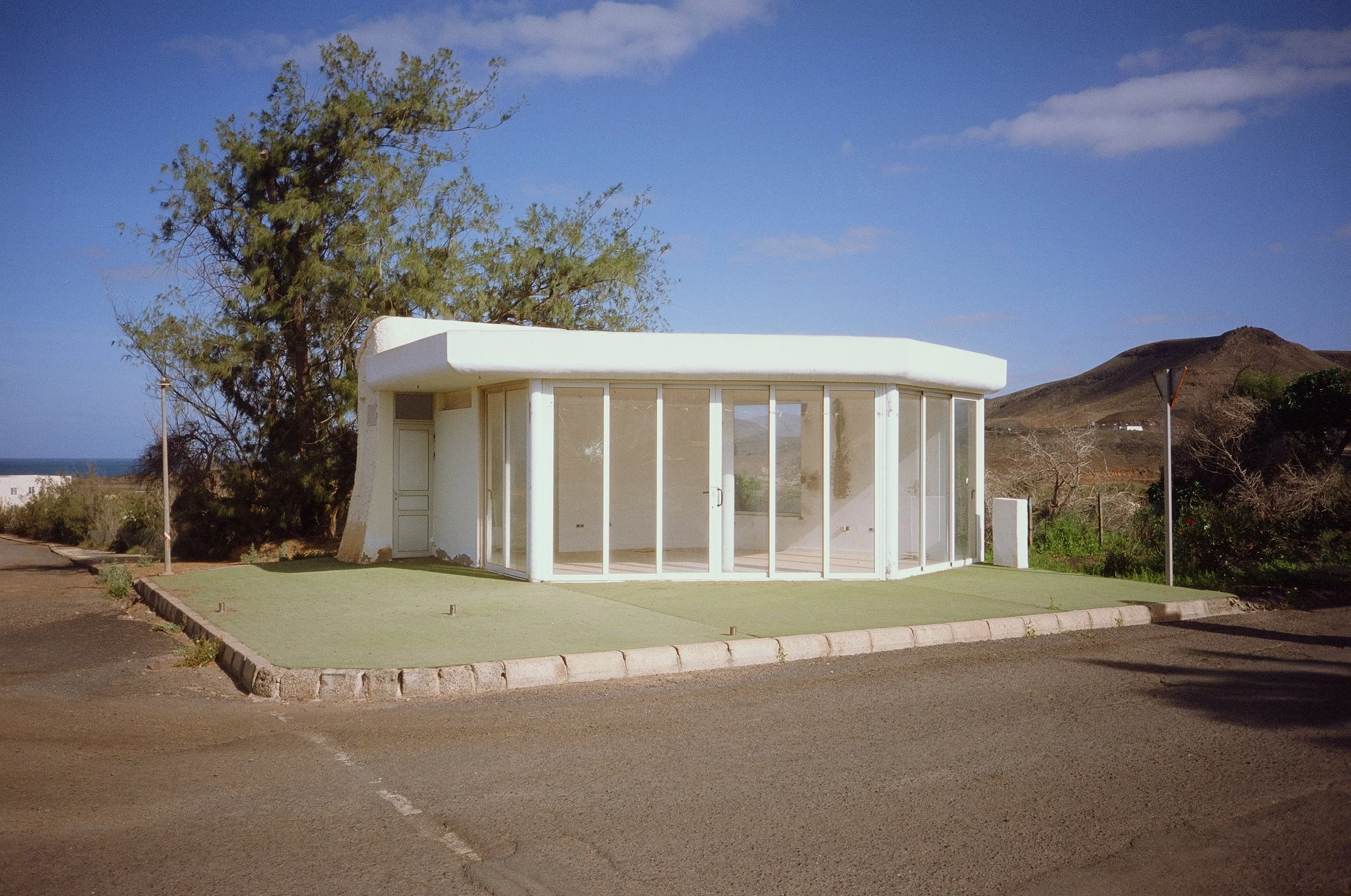 Small modern white building with large glass windows and a door, surrounded by a paved walk and a grassy area, with trees and hills in the background under a blue sky.