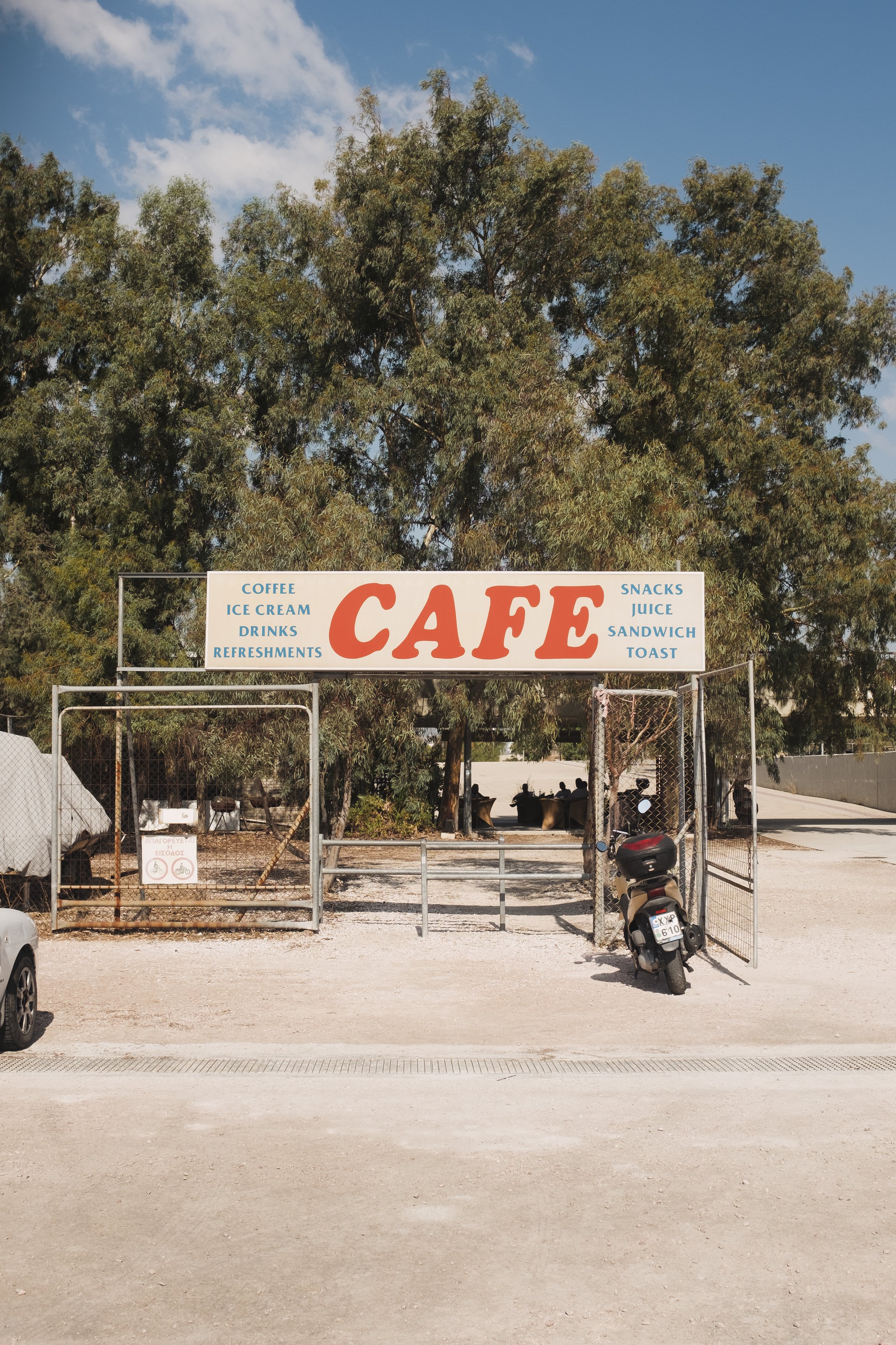 Entrance to a rustic cafe with a large sign featuring the word 'CAFE' and additional offerings like coffee, ice cream, drinks, refreshments, snacks, juice, sandwich, and toast, surrounded by trees and a few parked vehicles.
